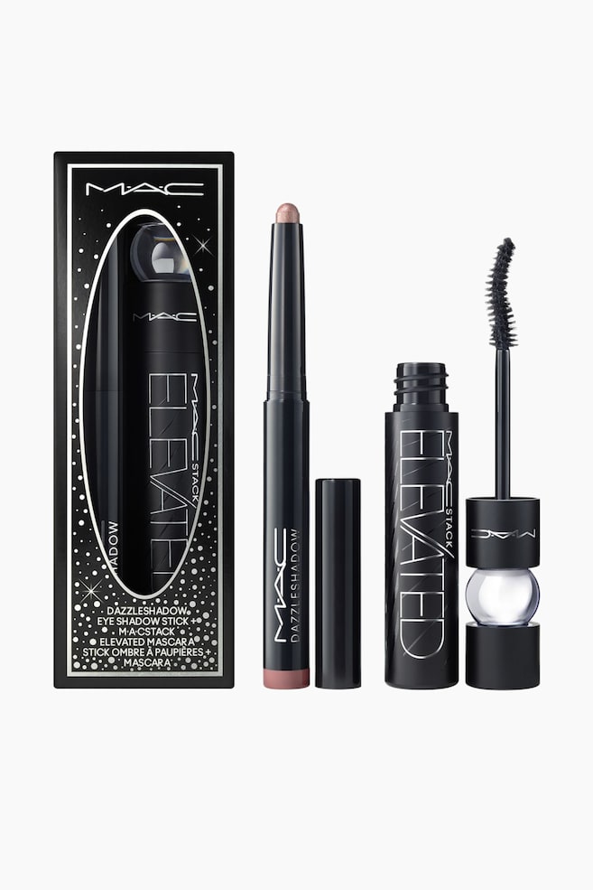 Dressed To Dazzle Eye Duo presenterer komponentene sine sammen med en svart og sÃ¸lvfarget eske med stjernemÃ¸nster. Oppreist stÃ¥r en Ã¥pen, skimrende Ã¸yenskyggepenn i champagnefarge med hetten tatt av, og en Ã¥pen svart mascara-tube med den buede bÃ¸rstapplikatoren plassert vertikalt ved siden av. - 1