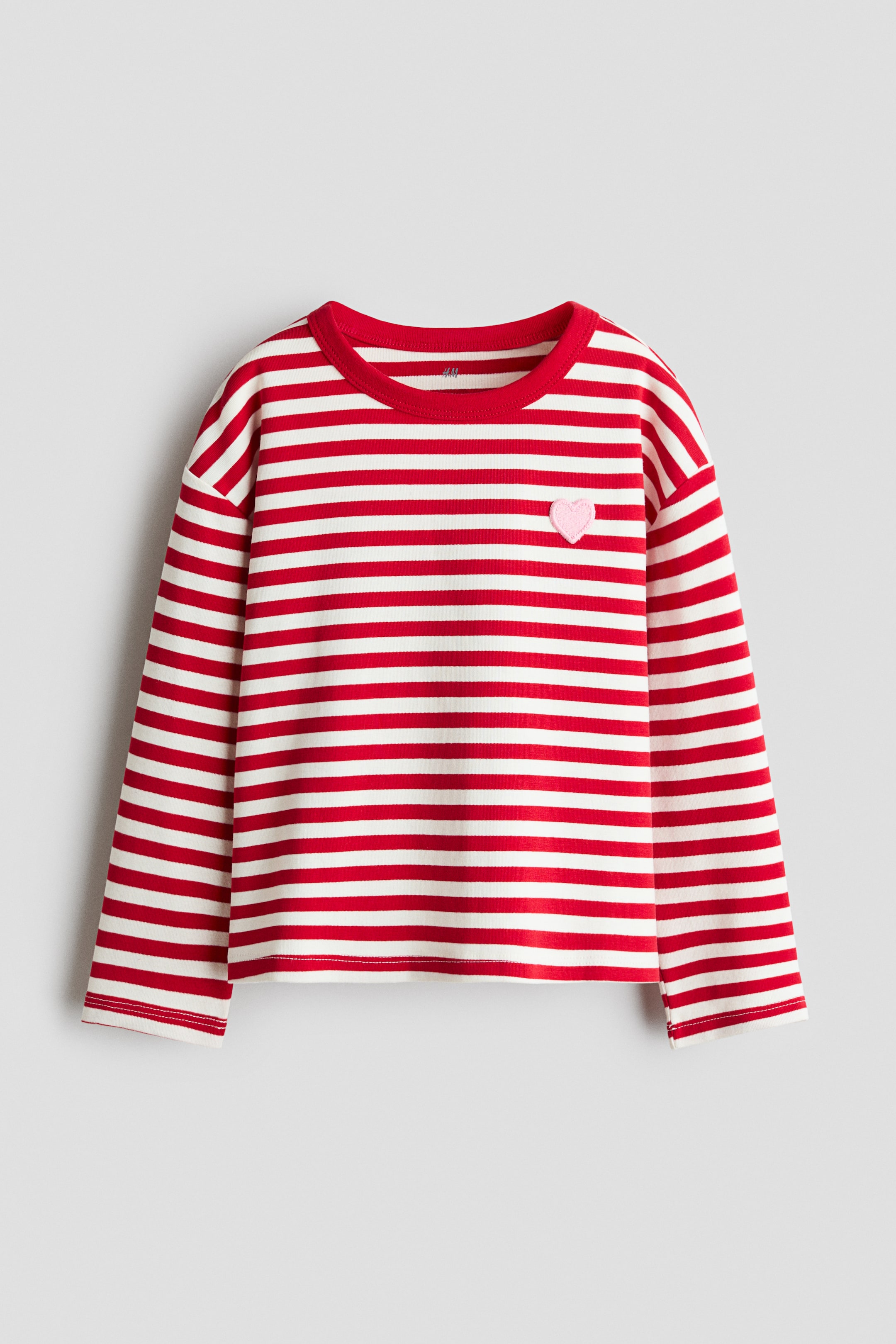 View larger image: A long-sleeved shirt features red and white horizontal stripes, a red round neckline, dropped shoulders, and a pink embroidered heart emblem on the upper left chest.