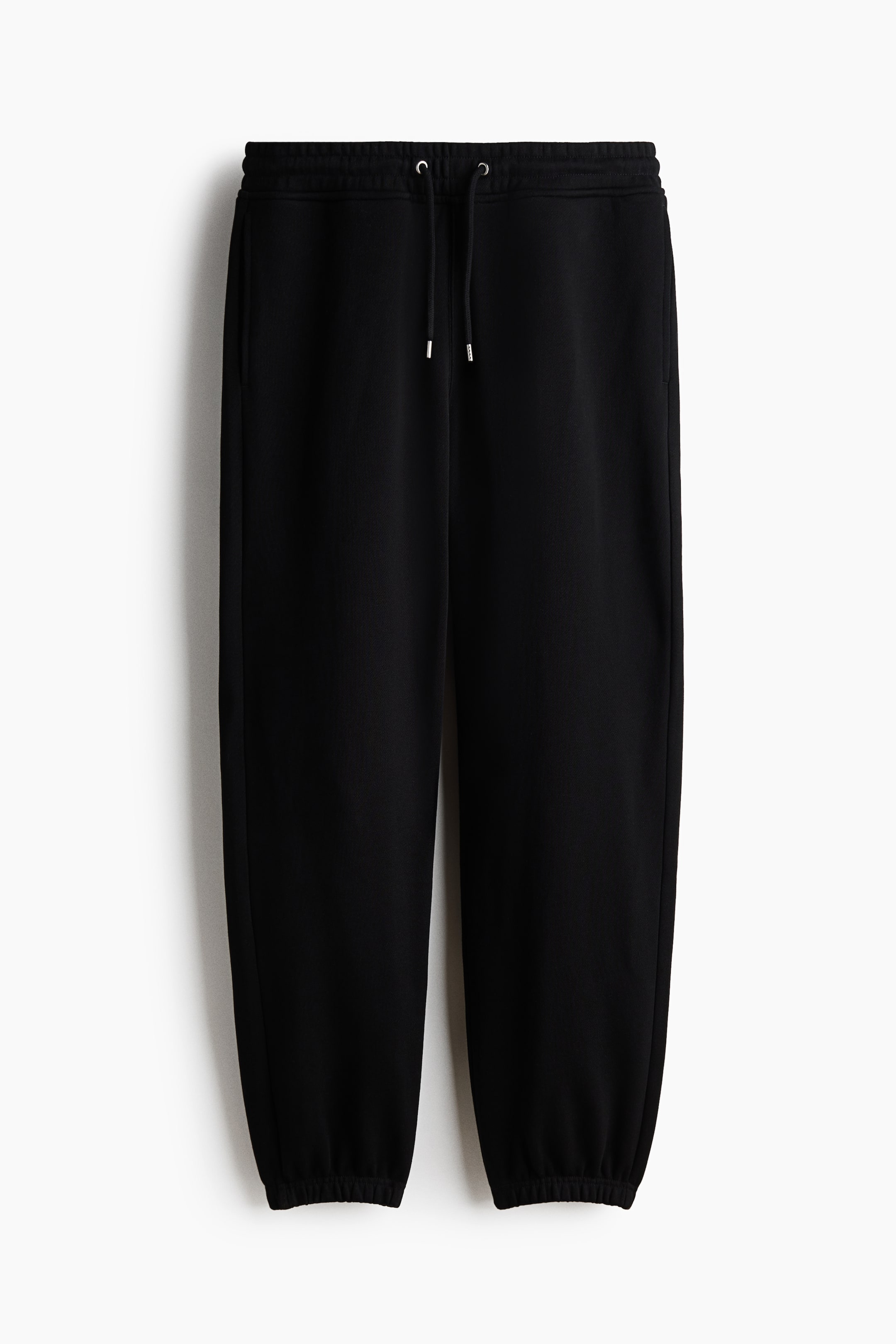 查看大圖: 黑色運動褲採用棉質運動衫面料製成，正面展示，設有覆蓋的彈性腰帶，配有抽繩和金屬鞋帶頭。這款運動褲呈現出寬鬆的版型，配有隱約的側縫口袋和彈性褲腳。