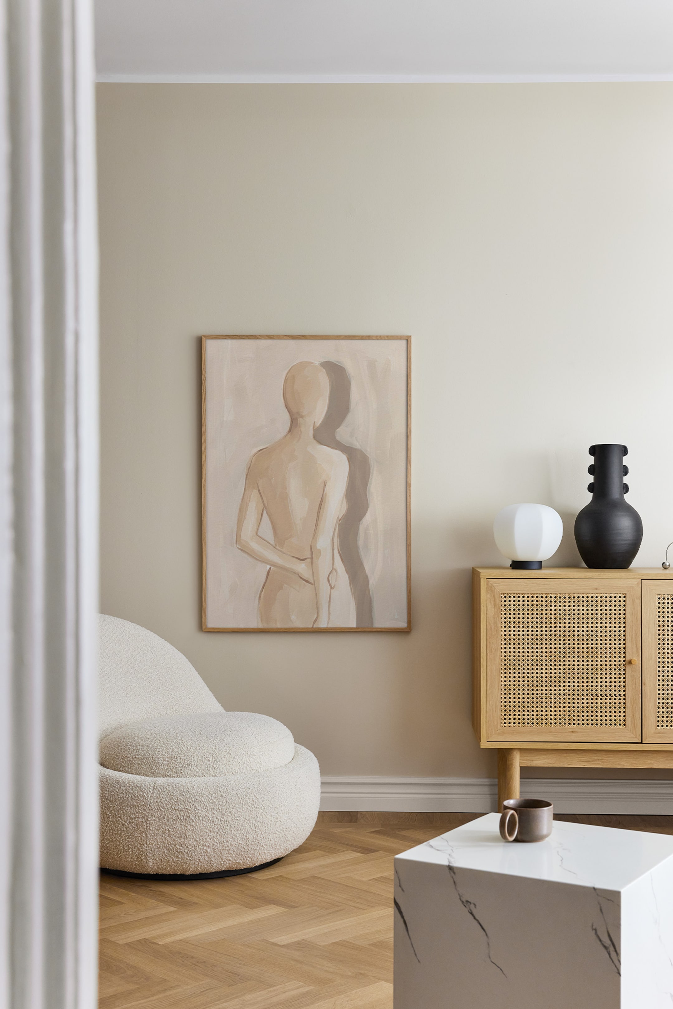 Grotere afbeelding bekijken: Een rechthoekige ingelijste kunstprint hangt vlak tegen een neutrale muur en toont een minimalistisch, abstract schilderij van een figuur van achteren, met een zachte schaduw die naar de zijkant valt. Het kunstwerk bevat warme beige en grijze penseelstreken en is omlijst met een lichtgekleurde houten lijst.