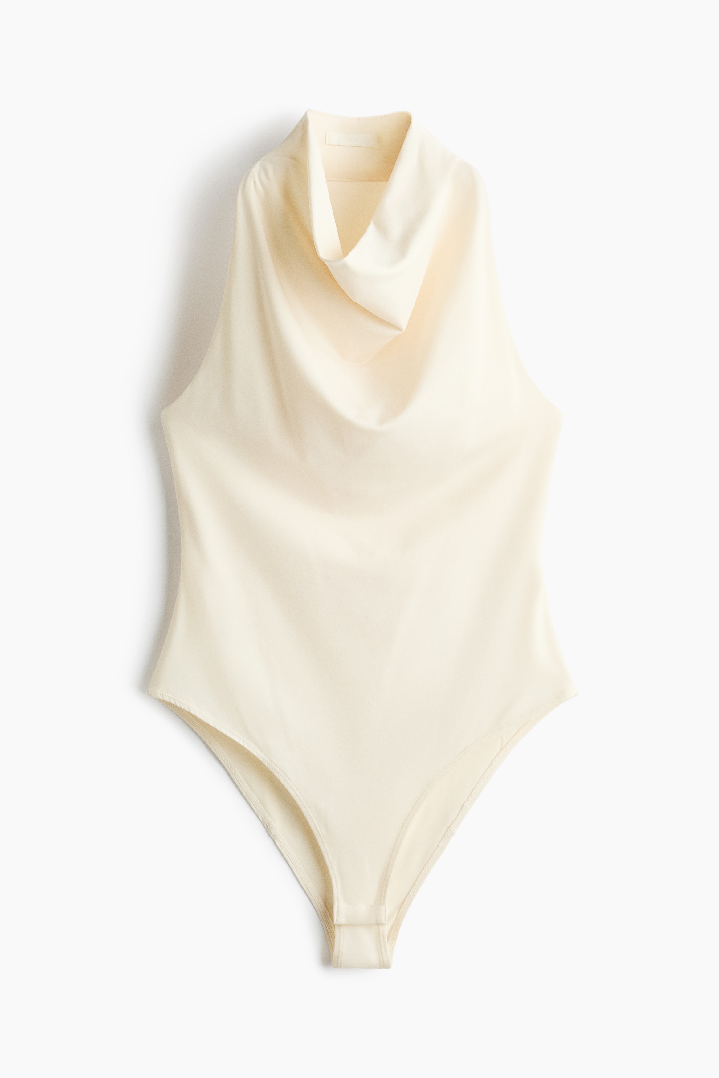 La modelo lleva puesto un bodysuit de color claro, sin mangas, con cuello drapeado tipo cowl y un corte estrecho en los hombros. El bodysuit está metido dentro de unos pantalones de color claro, y la modelo sostiene las mangas de un suéter amarrado a la cintura.