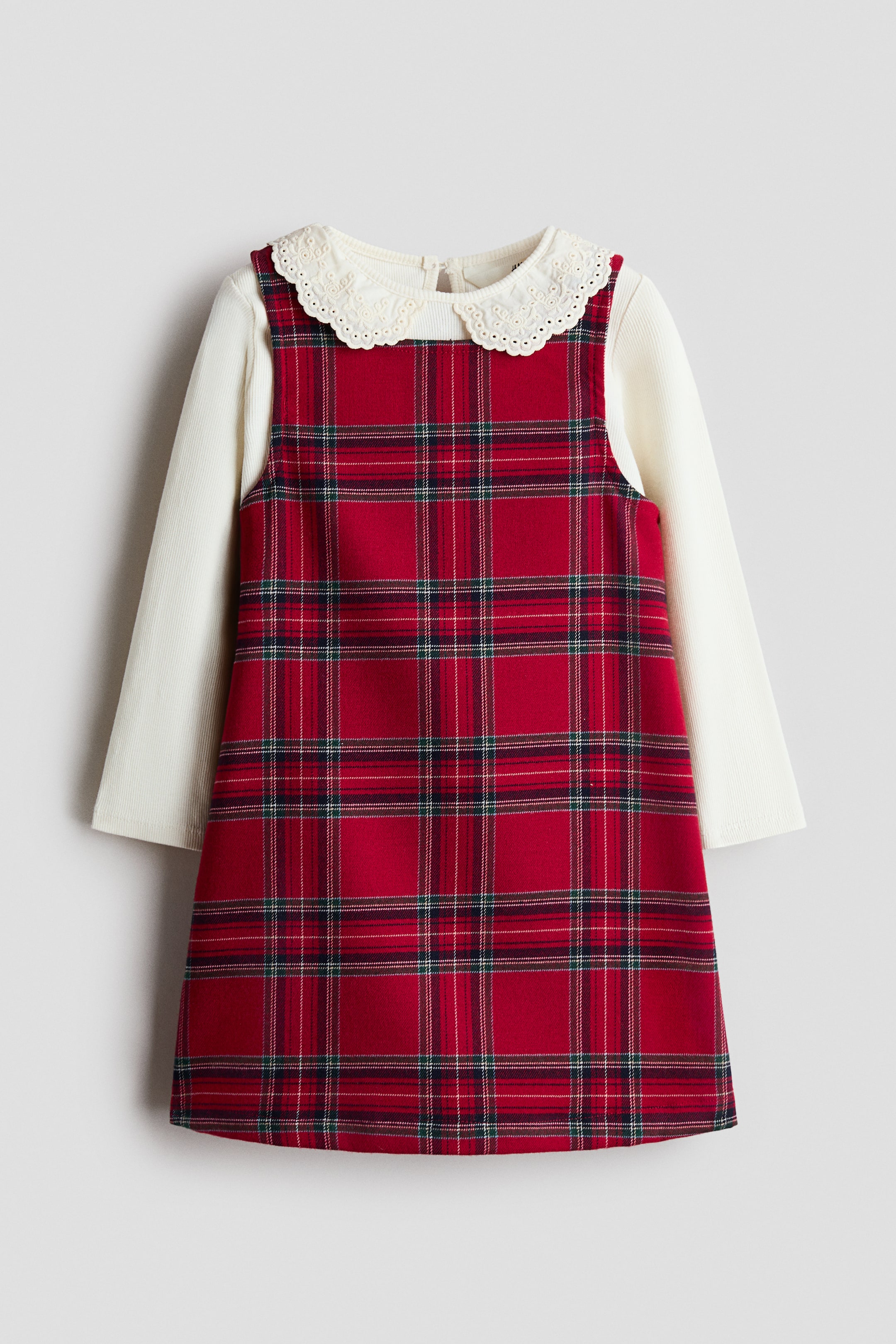 View larger image: A red plaid pinafore dress is layered over a cream-coloured, long-sleeved top, shown from the front. The sleeveless dress features a red, green, and white tartan pattern, while the top displays a ribbed texture and a white, eyelet-embroidered Peter Pan collar.