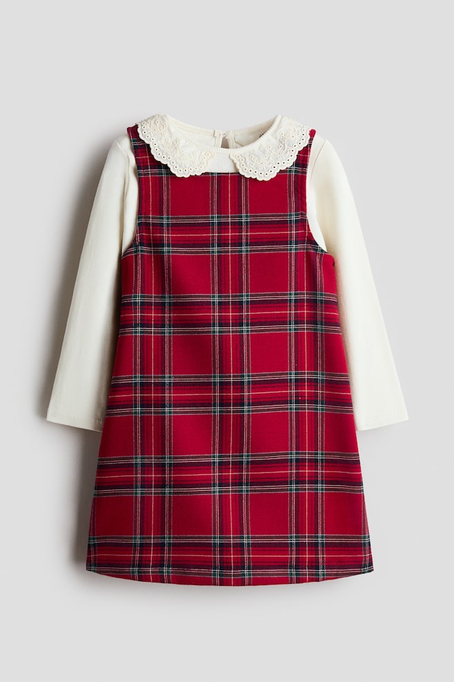A red plaid pinafore dress is layered over a cream-coloured, long-sleeved top, shown from the front. The sleeveless dress features a red, green, and white tartan pattern, while the top displays a ribbed texture and a white, eyelet-embroidered Peter Pan collar. - 1