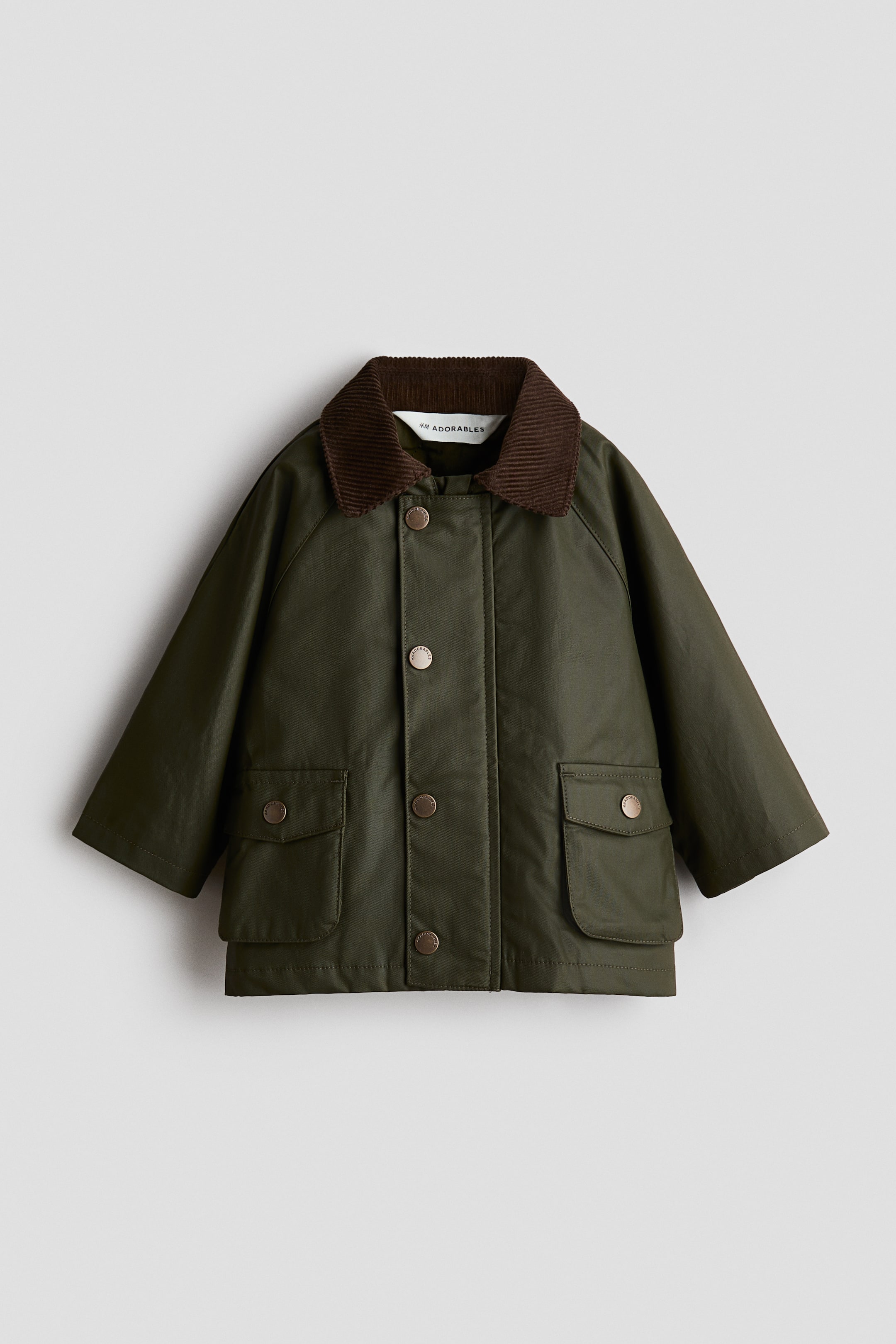 Visa större bild: H&M Adorables-jackan är vänd framåt och har ett olivgrönt vaxat twillmaterial med en brun manchesterkrage och raglanärmar. Den visar upp en frontslå med metalltryckknappar och två påsydda framfickor, var och en med ett lock med metalltryckknappsstängning.