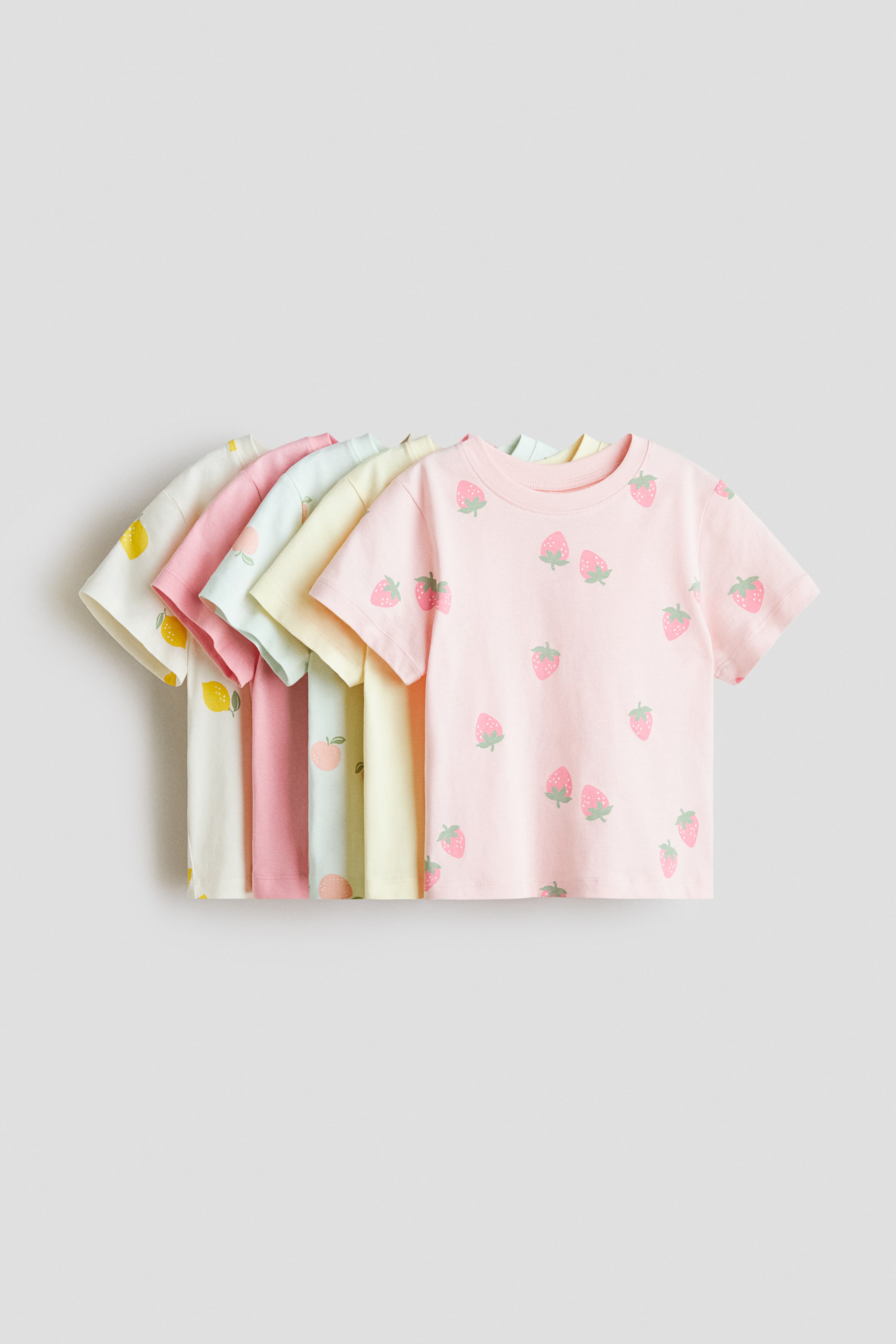View larger image: Five soft cotton jersey t-shirts are arranged front-facing, fanning out in a row. Each t-shirt is designed with short sleeves and a ribbed trim around the neckline, displaying fruit patterns of strawberries, peaches, and lemons on pastel-coloured fabrics.