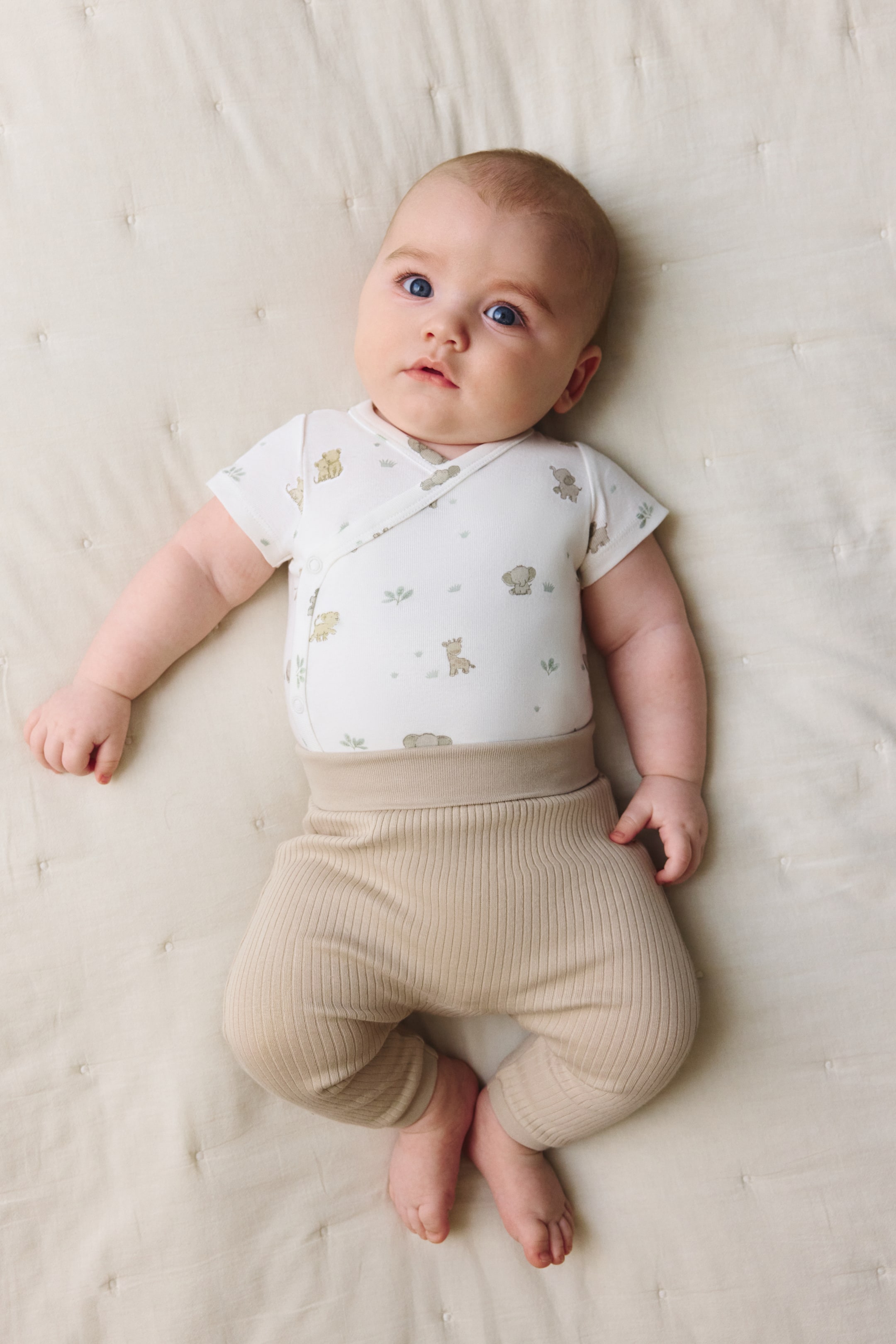 Ingrandisci l'immagine: Un body bianco a maniche corte in stile avvolgente, stampato con figure di animali dai colori chiari e fogliame verde, è abbinato a pantaloni beige a coste con un'ampia fascia elastica in vita e polsini a coste alle caviglie.