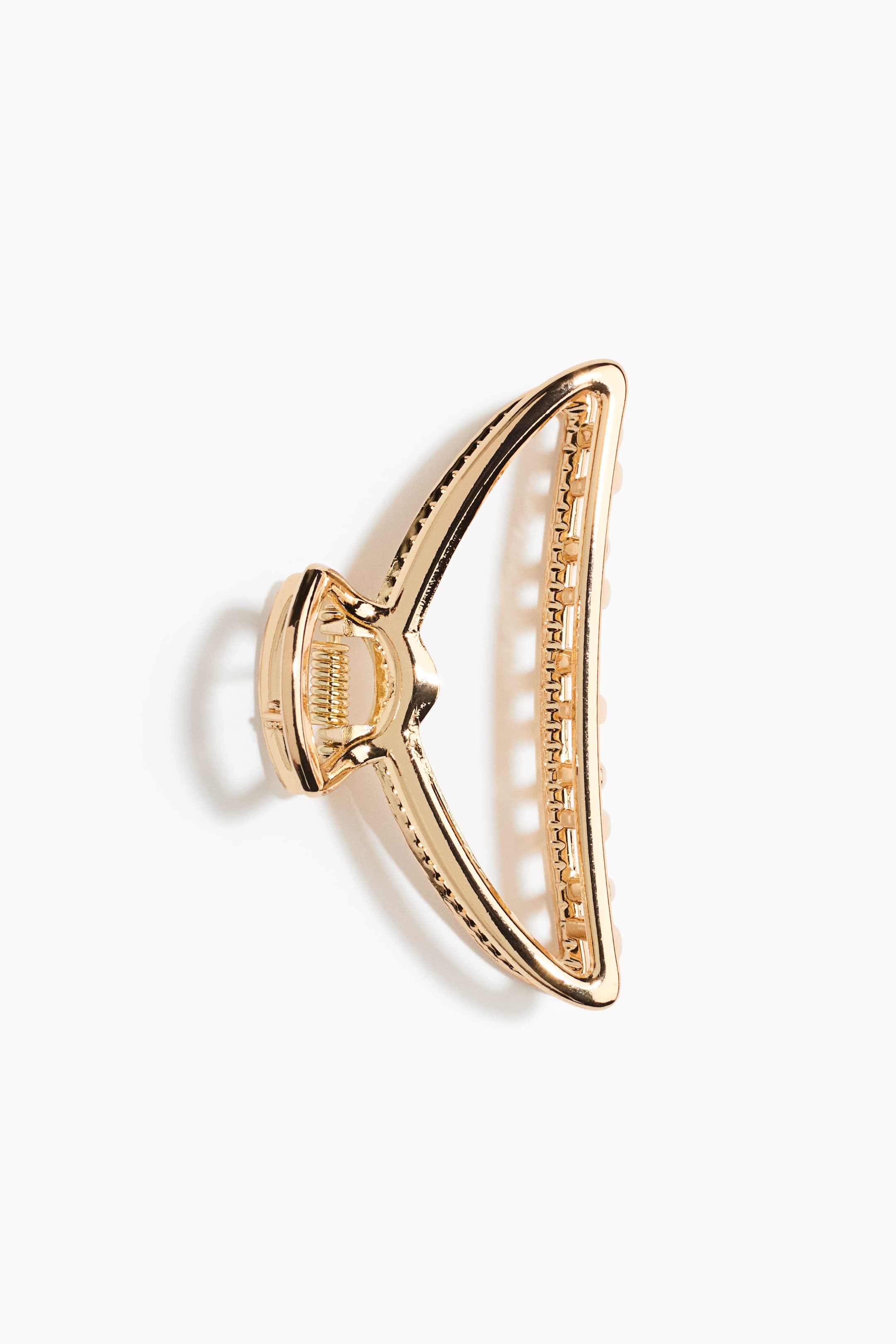 View larger image: A gold-toned metal hair claw clip is resting flat, showcasing its open-sided, curved triangular structure. The clip is displaying a polished metallic finish, a visible spring coil mechanism on one end, and interlocking teeth along the inner edge.
