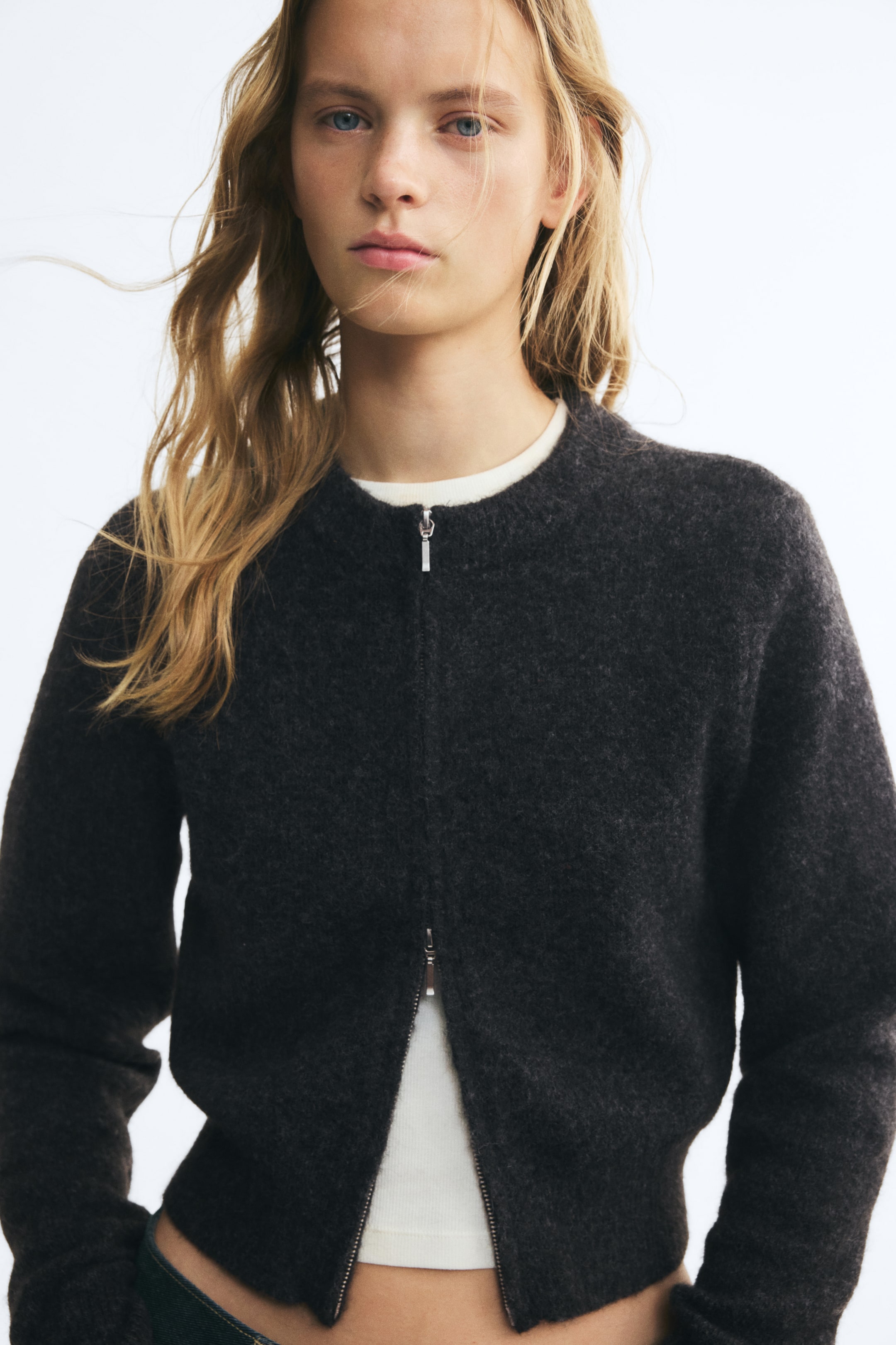 Grotere afbeelding bekijken: Een donkergrijze, zachtgebreide korte cardigan met een ronde geribde halslijn en lange mouwen wordt door het model gedragen over een witte top. De cardigan heeft een tweewegrits aan de voorkant, die bij de hals gesloten is en bij de geribde zoom open.