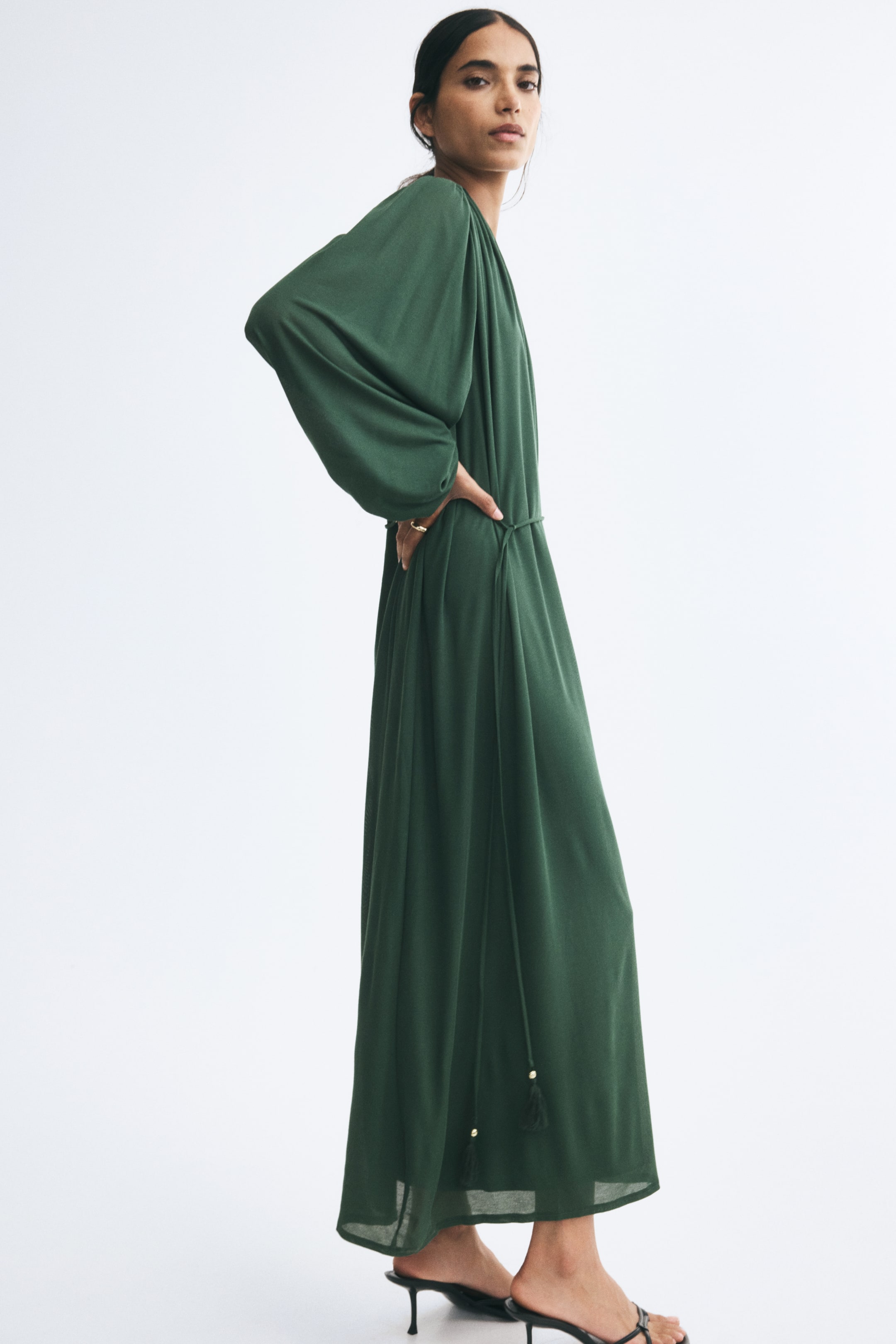 View larger image: A long, loose-fit dark green dress in fine ribbed jersey is being worn by the model, who is standing in profile with one hand resting on her hip. The dress features long balloon sleeves with elasticated cuffs and a thin tie belt at the waist with tasselled, beaded ends.