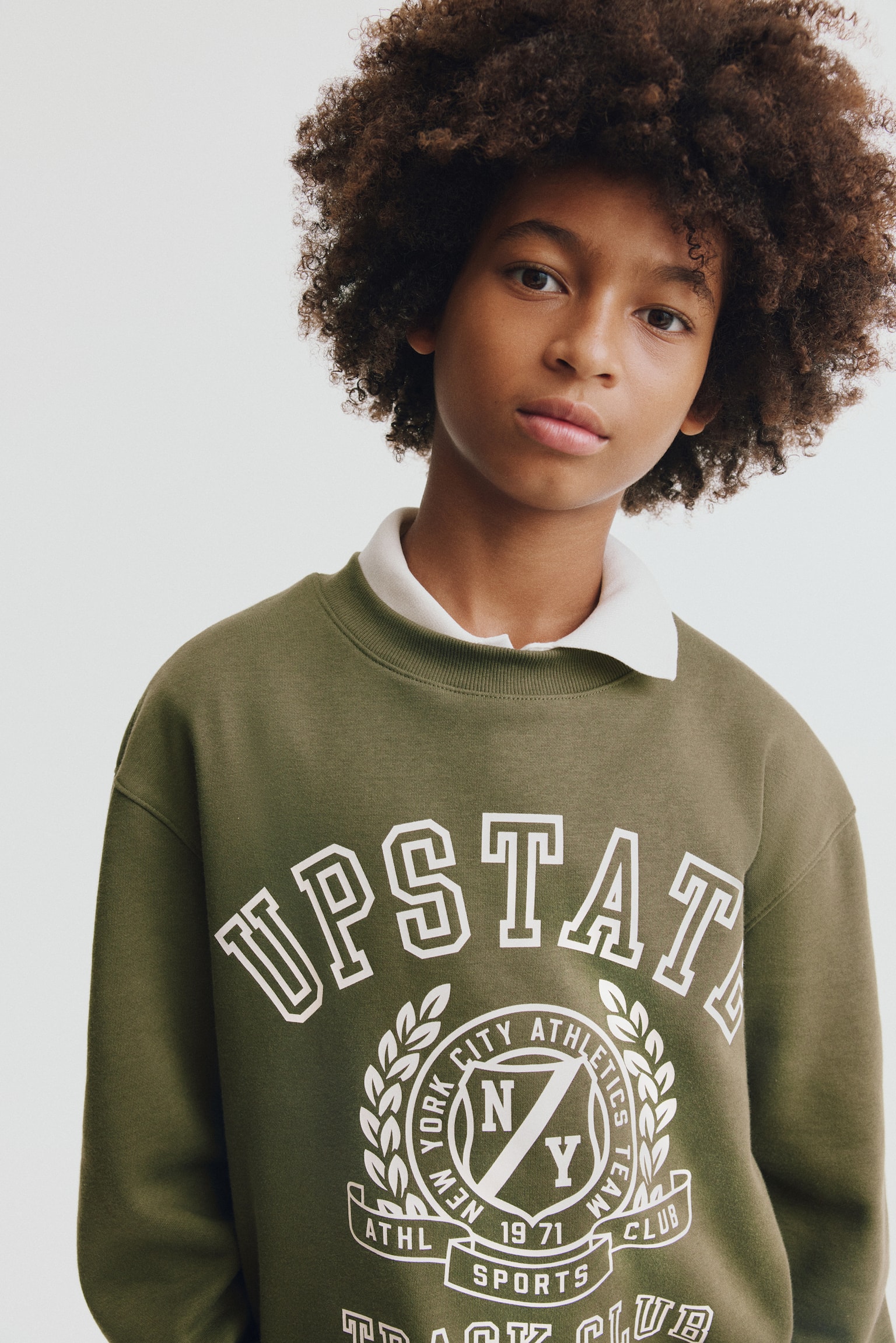 An olive green sweatshirt is shown from the front, featuring a round, rib-trimmed neckline, dropped shoulders, and a white collegiate-style graphic on the chest reading "UPSTATE" above an athletic crest. The sweatshirt is layered over a white collared shirt, with the collar peeking out from the neckline. - 1