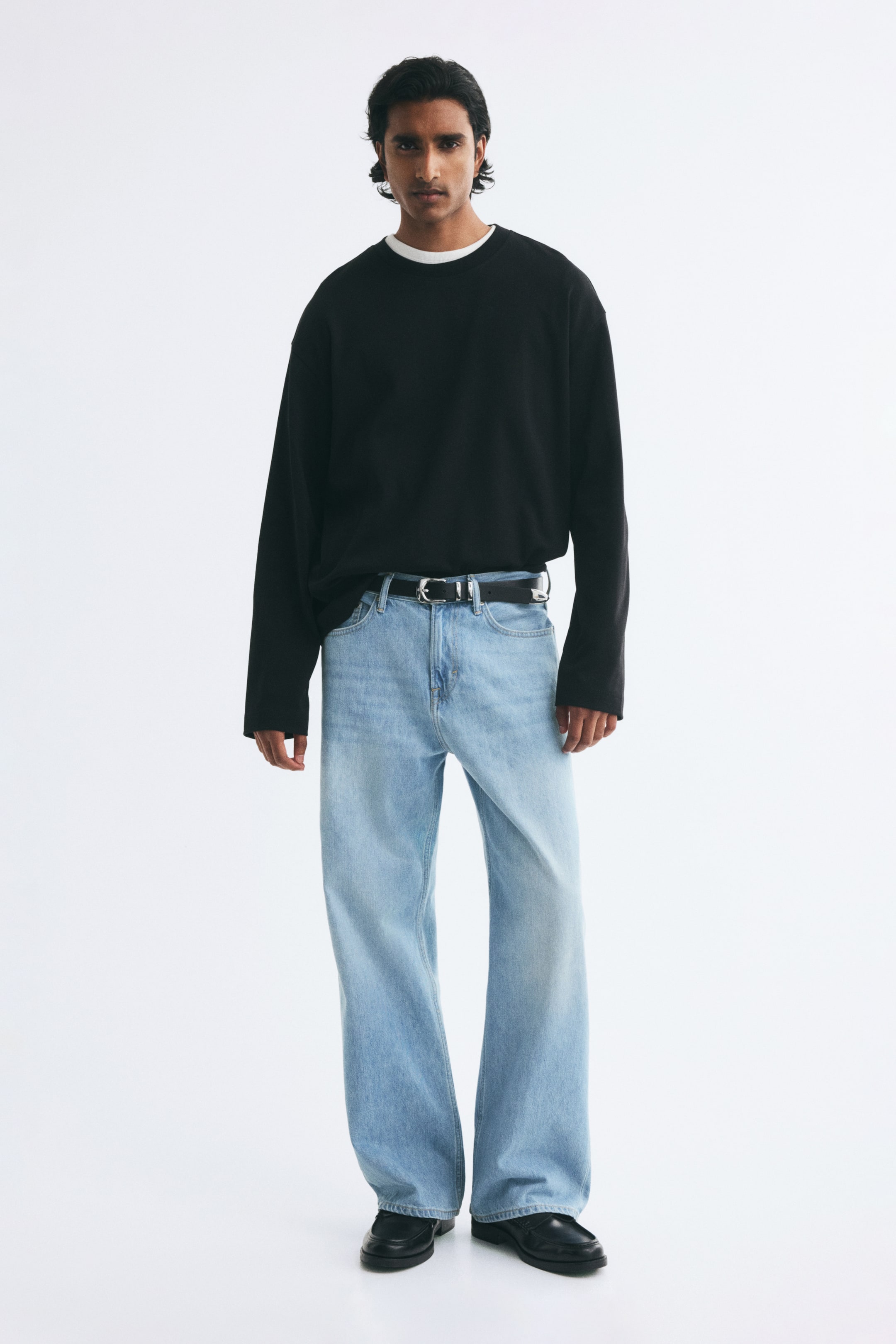 View larger image: The model is wearing light blue loose-fit denim jeans featuring a regular waist, a dropped crotch, and wide straight legs. The jeans are styled with a black leather belt and a black long-sleeved shirt tucked into the waistband.