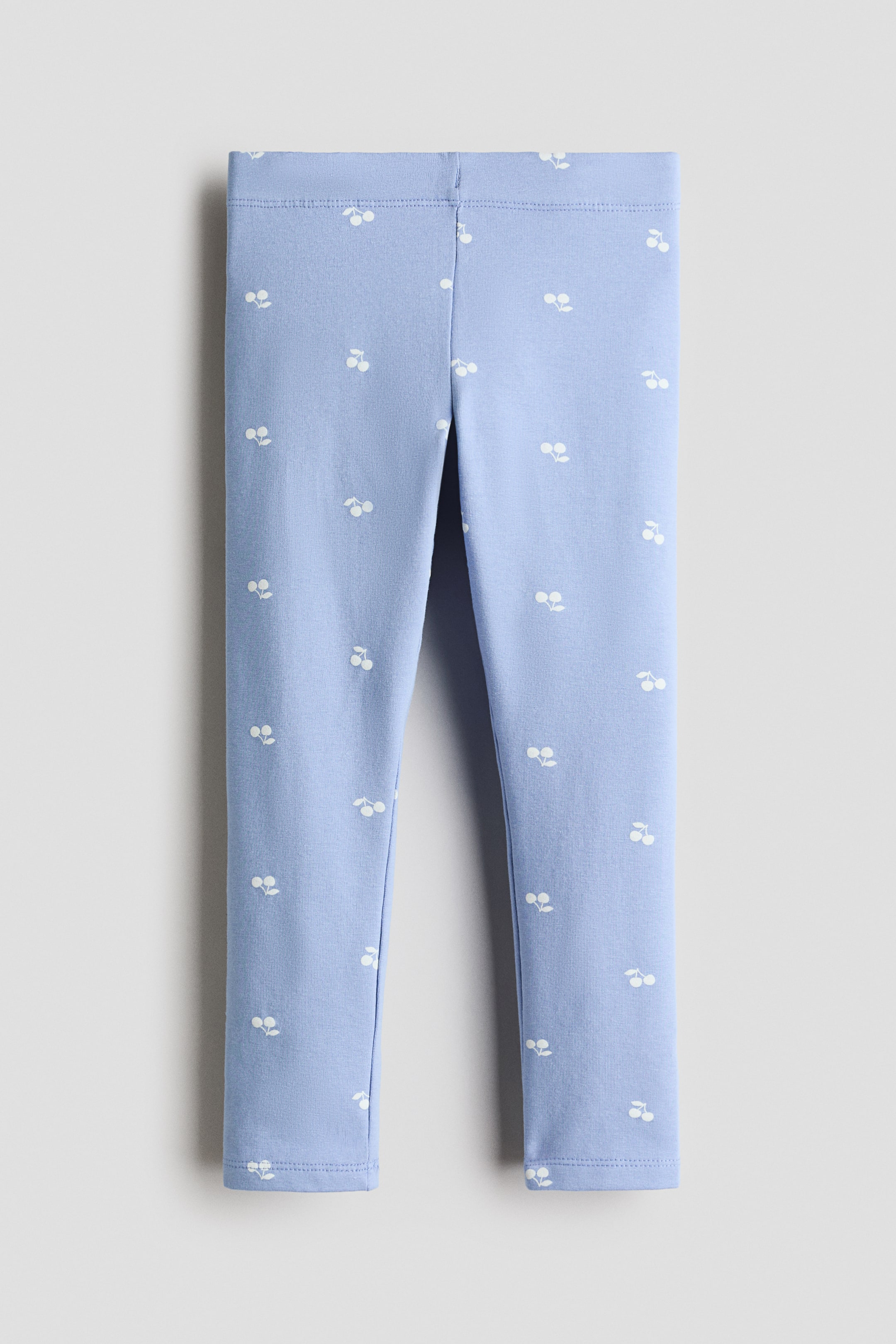 View larger image: Light blue leggings are laid flat, featuring an all-over pattern of small white cherry motifs, a flat elasticated waistband, and simple stitched ankle hems.