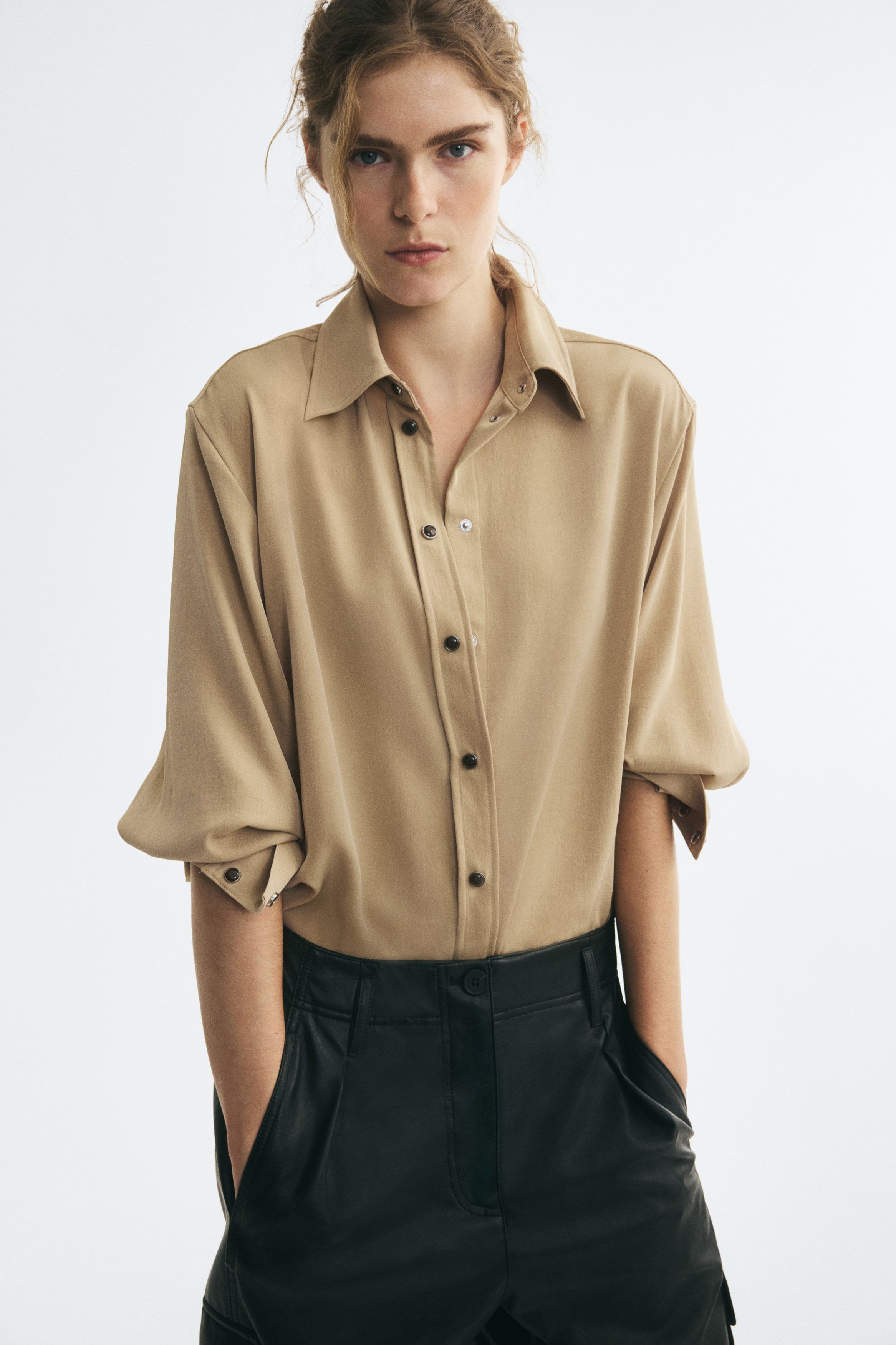 An oversized beige shirt in a woven fabric is shown from the front, featuring a pointed collar, dropped shoulders, and a front placket with dark marbled buttons. The shirt is designed with long balloon sleeves ending in wide cuffs and a gently rounded hem.