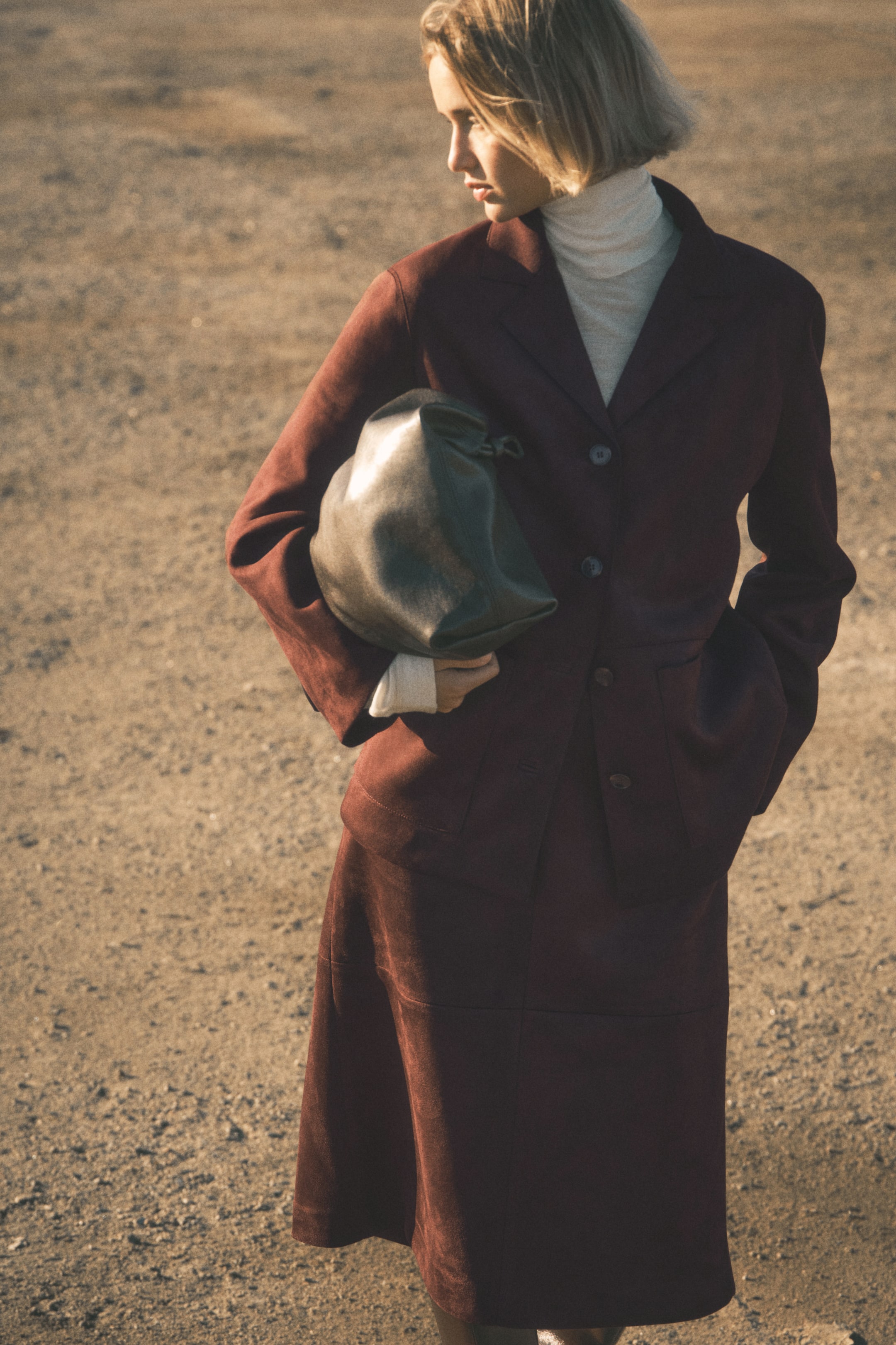 Vezi imaginea mai mare: O jachetă din piele întoarsă de culoare maro-roșcat închis și o fustă midi asortată sunt prezentate dintr-un unghi ușor frontal-stânga. Jacheta prezintă reveruri cravate, două nasturi și două buzunare aplicate, fiind purtată peste un pulover cu guler înalt de culoare deschisă. Fusta midi are un detaliu de cusătură orizontală în jurul șoldurilor, iar o geantă șleampătă din piele gri închis cu închidere cu șnur este ținută sub braț.