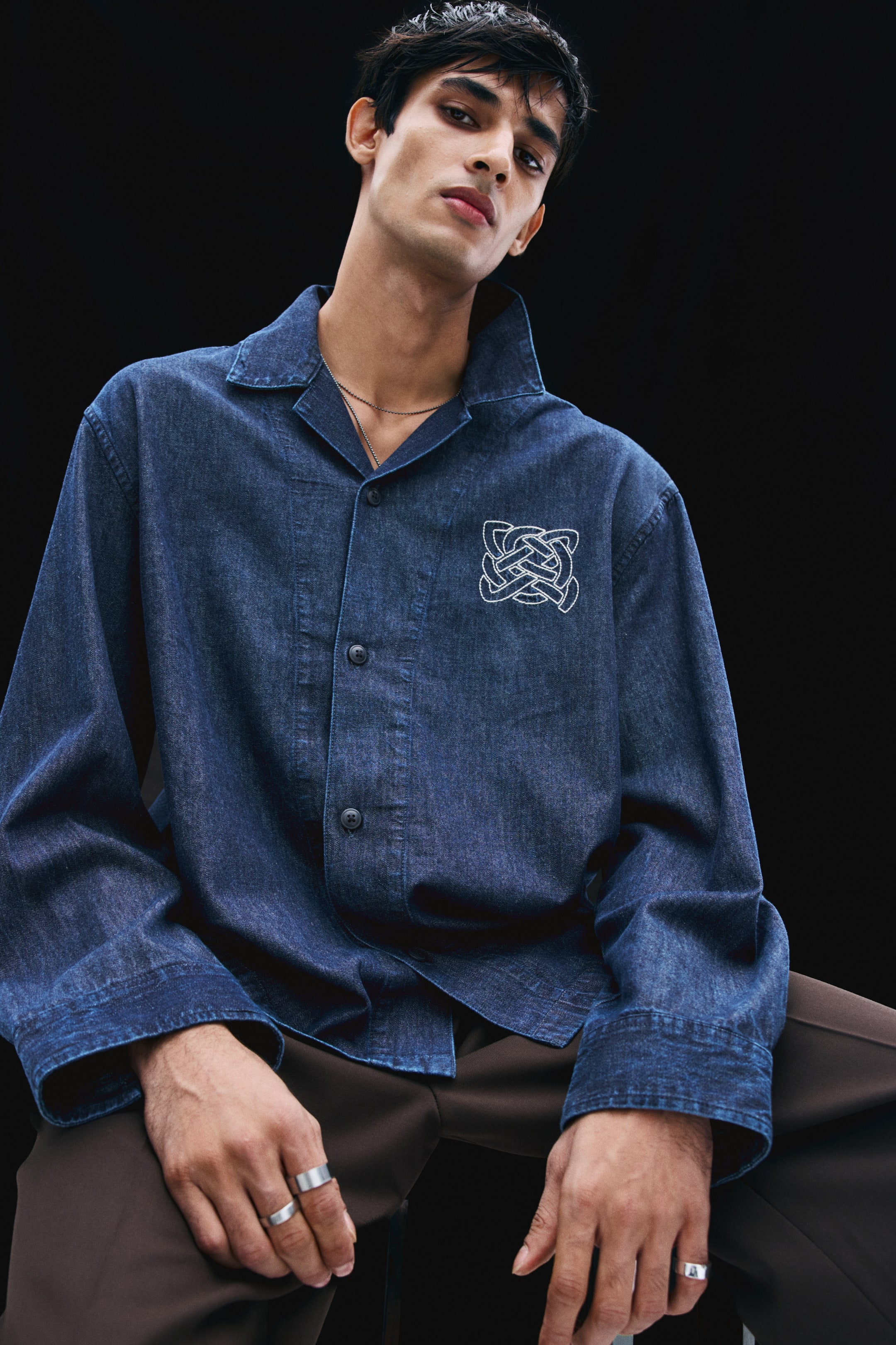 View larger image: A dark blue denim overshirt is worn by the model, featuring a resort collar, a French front with button closures, and a white embroidered knot design on the left chest. The shirt is styled with a thin chain necklace and paired with brown trousers.