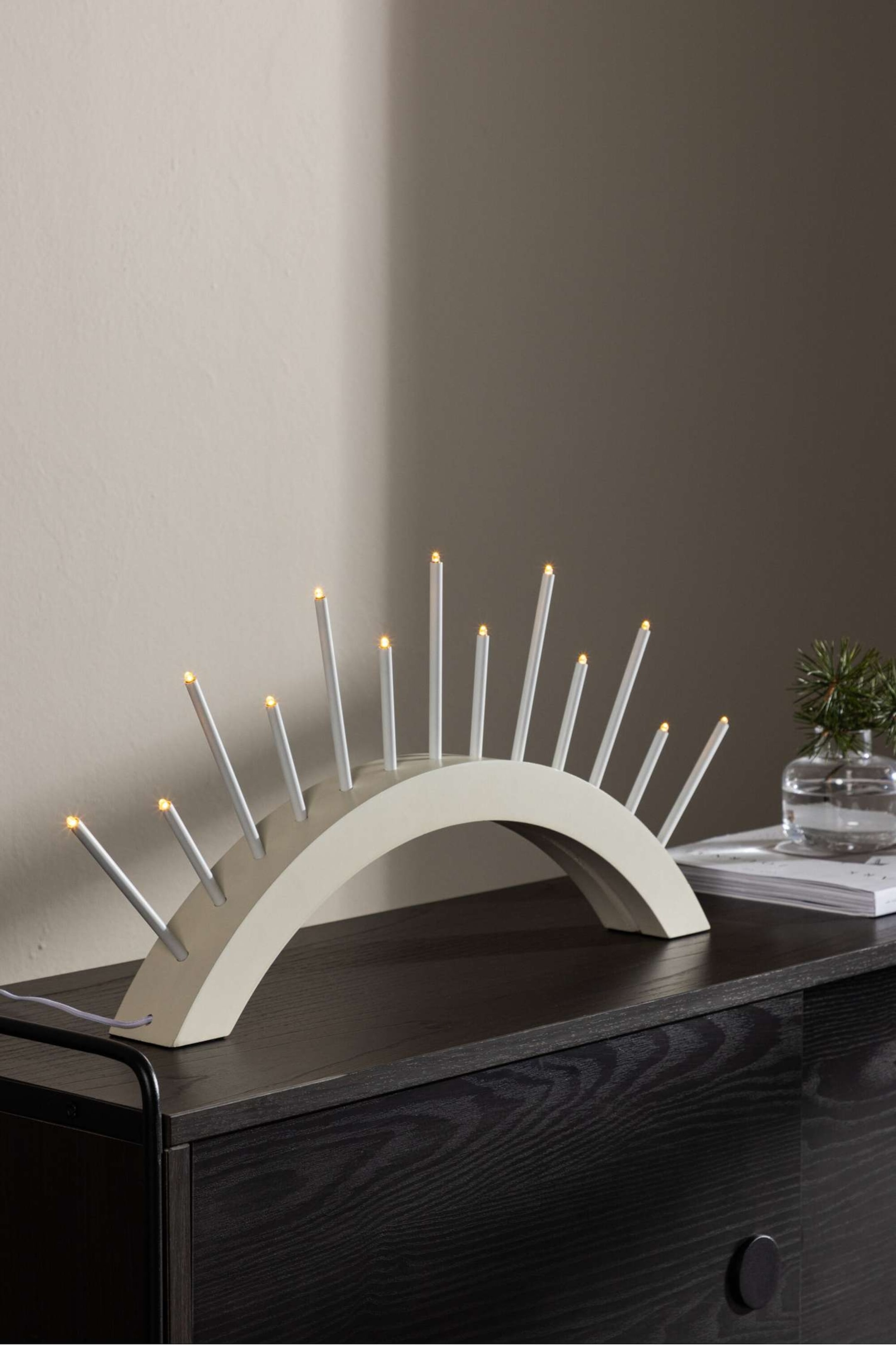 Agrandir l'image: Un candélabre électrique arqué doté d’une base semi-circulaire lisse, blanc cassé, avec onze fines bougies blanches de hauteurs variées, disposées en éventail vers l’extérieur. Chaque bougie est surmontée d’une petite lumière LED chaleureuse et lumineuse évoquant une flamme.