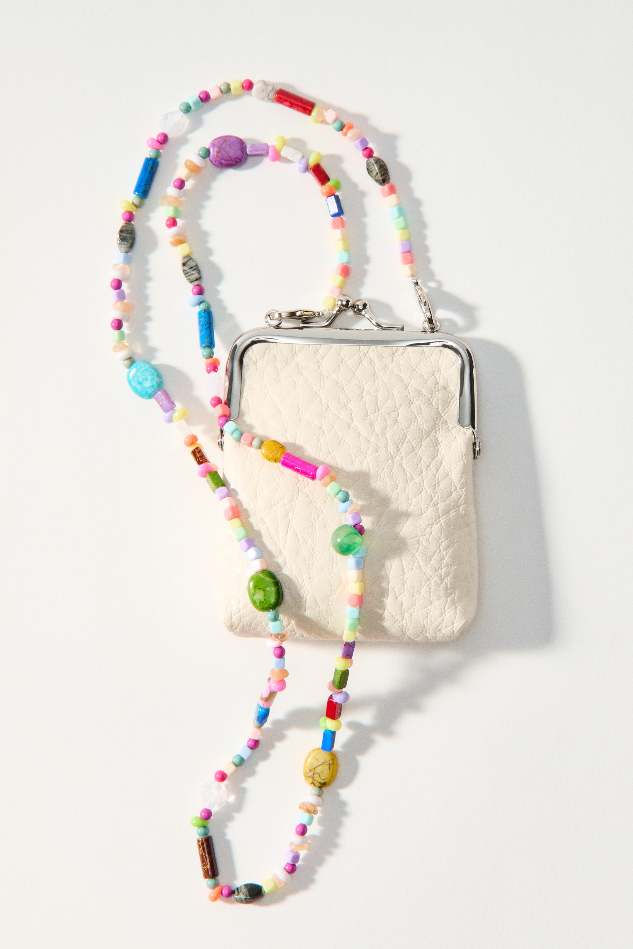 Se større billede: En lille crossbody-taske vender fremad og har et off-white belagt stofkorpus med en sølvfarvet metalramme og en pungelukning øverst. En aftagelig skulderrem, dekoreret med flerfarvede plastikperler i forskellige former og størrelser, er draperet omkring tasken.