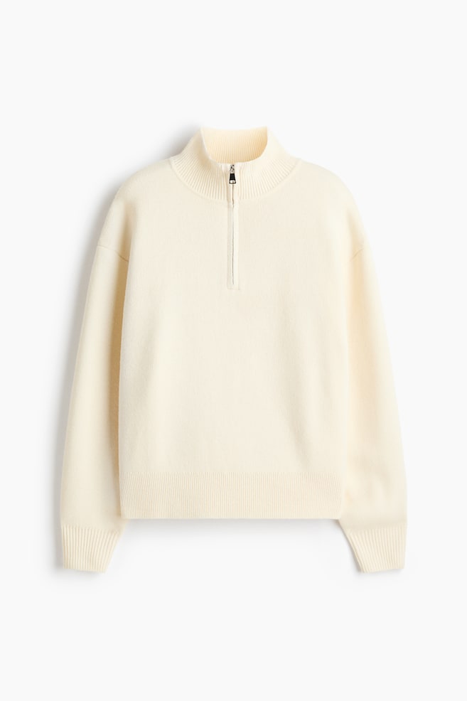 An oversized cream-colored jumper is shown from the front, featuring a fine-knit texture, a rib-trimmed turtleneck with a partial zipper, and dropped shoulders. The design is showcasing long sleeves with ribbed cuffs and a matching ribbed hem. - 1