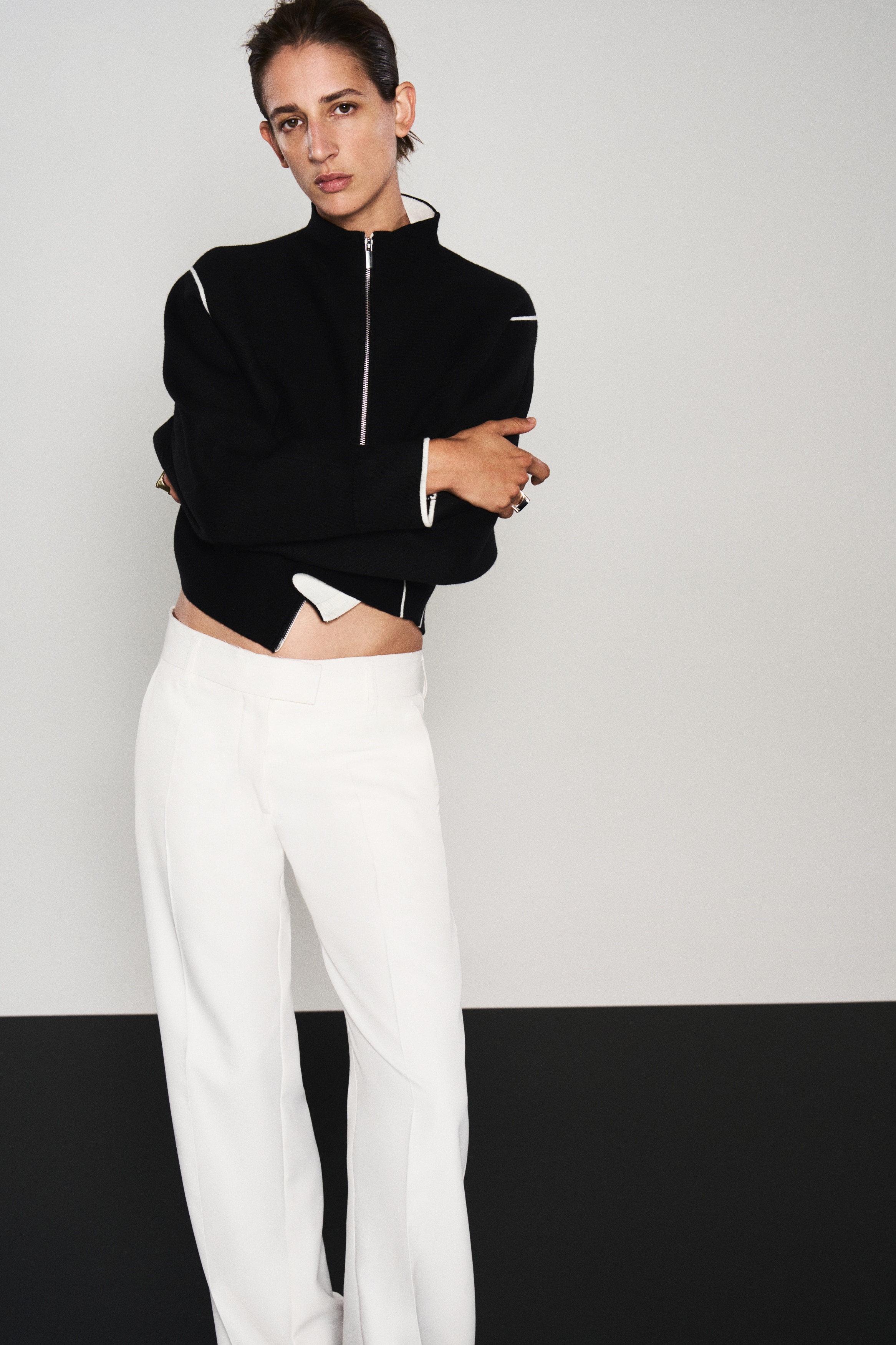 A black knit zip-up jacket is shown from the front, featuring a high stand collar with a contrasting white interior and a full-length silver zip. It has relaxed, wide sleeves defined by thin white piping along the shoulder and side seams, finished with a subtle white trim at the cuffs.