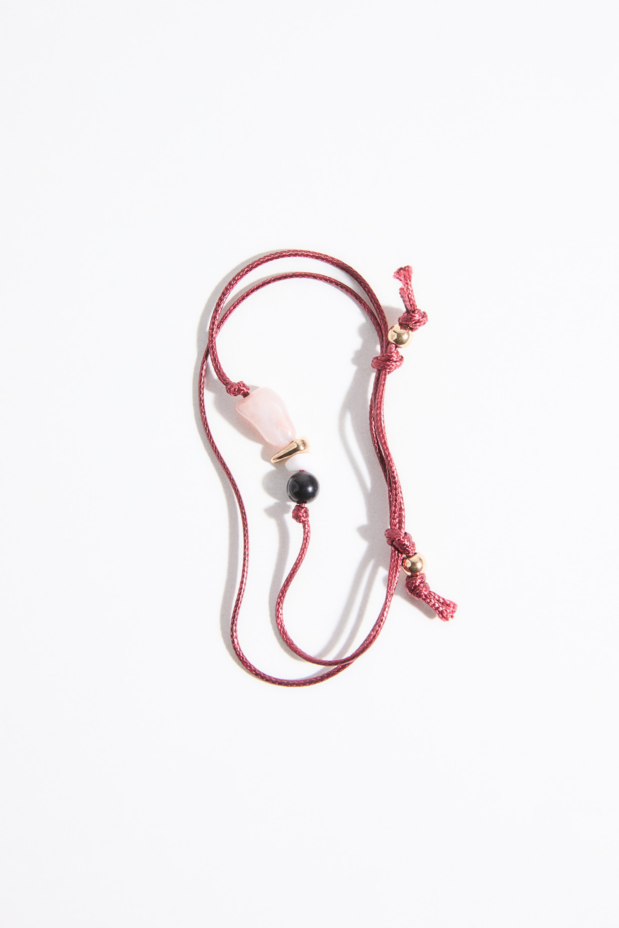 拡大画像を表示: マルーンのコードアクセサリーが緩やかなSカーブを描いて平らに置かれており、中心には淡いピンクの不規則な形状の石ビーズ、小さなゴールドのチューブスペーサー、そして黒い丸いビーズが配置されています。コードの両端には調整可能なスライドノットがあり、それぞれに小さなゴールドビーズが固定されています。