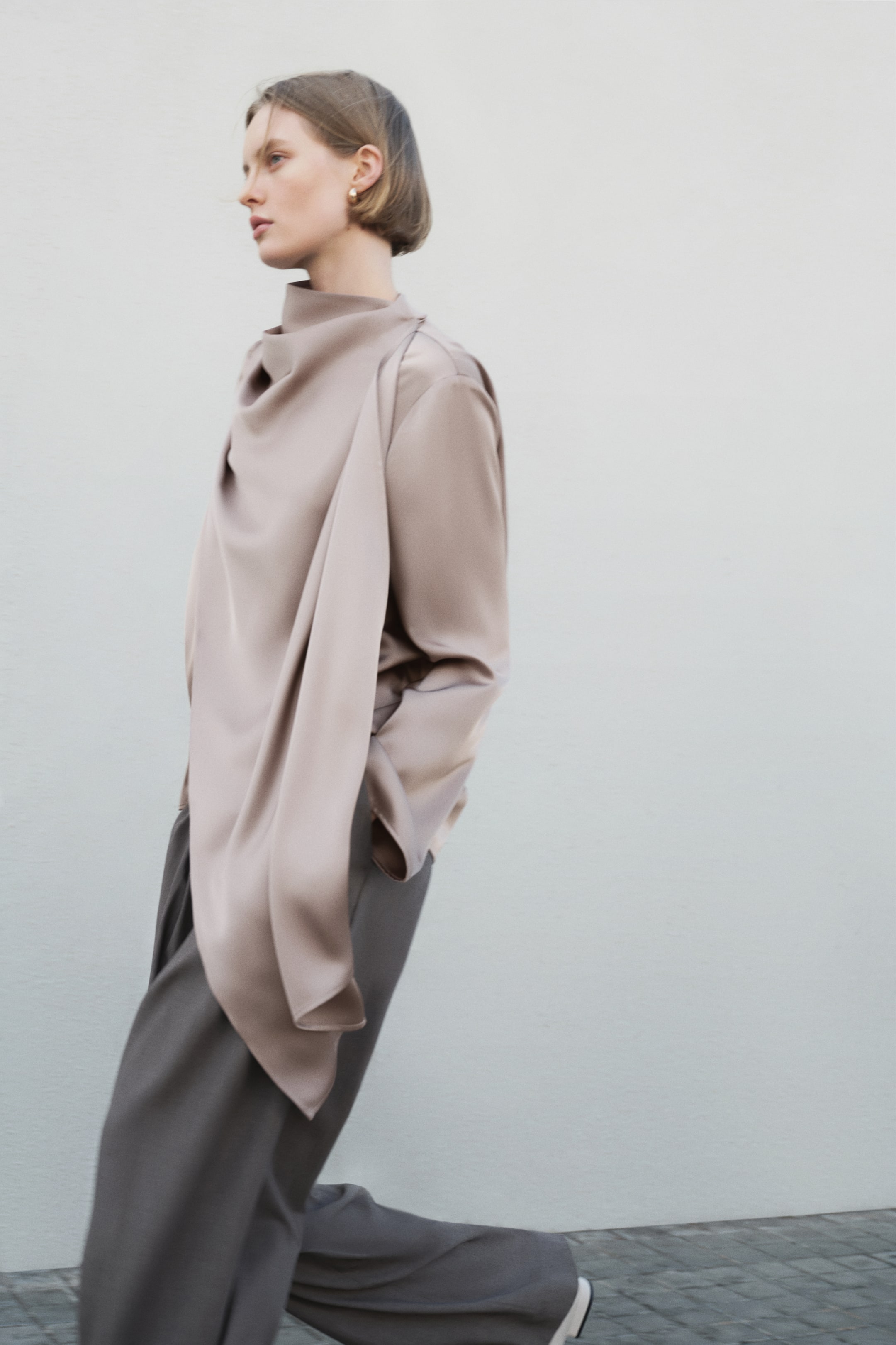 Xem ảnh lớn hơn: Một chiếc áo blouse satin màu be nhạt nổi bật với cổ áo không đối xứng, tạo thành một cổ cao và đổ xuống phía trước. Nó được kết hợp với quần tây rộng màu xám đậm.