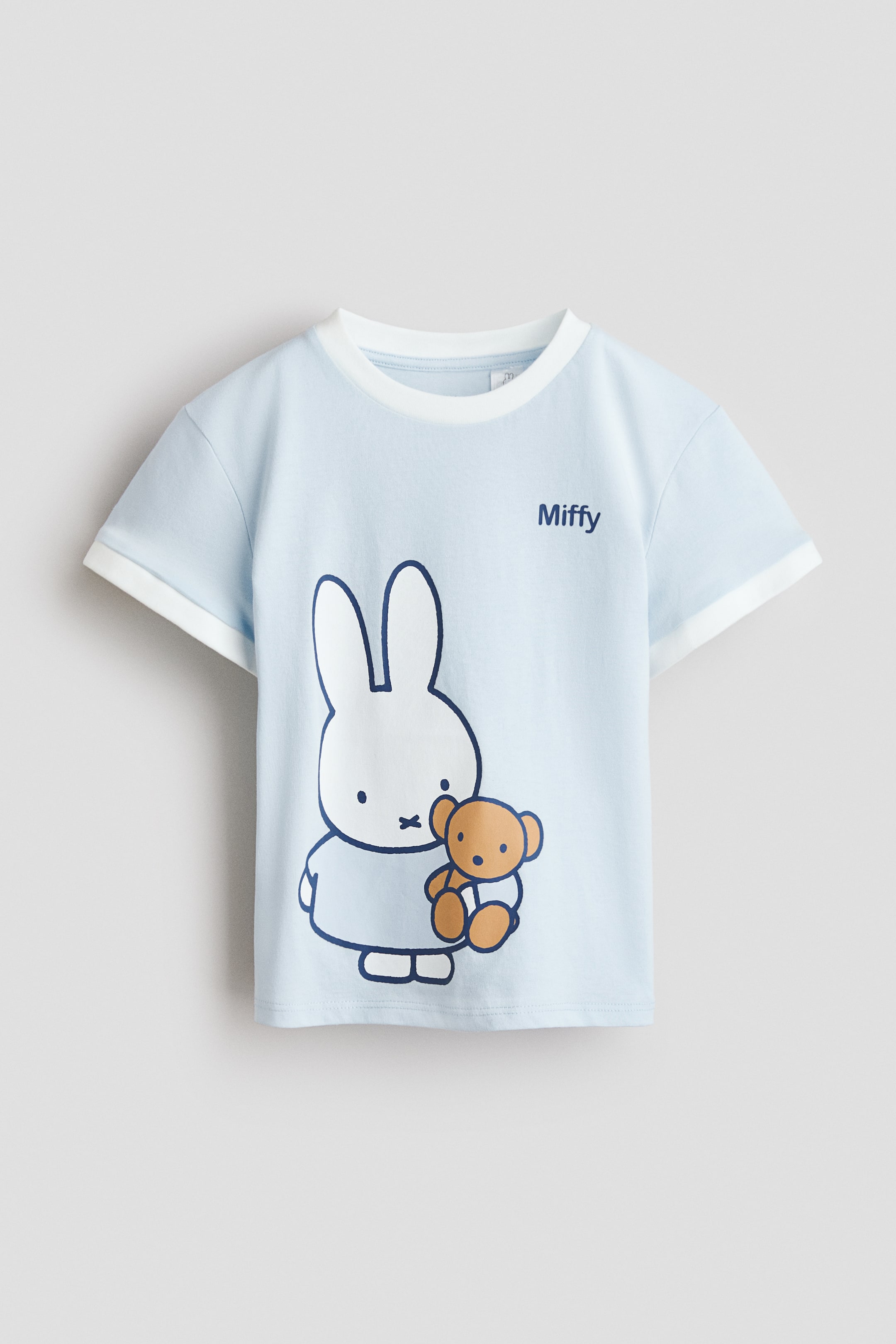 拡大画像を表示: ライトブルーのTシャツが前を向いており、白いクルーネックと白いトリムのショートスリーブが特徴です。前面には、濃いアウトラインで描かれた白いウサギのキャラクターが茶色のテディベアを抱えており、その上には「Miffy」という文字が濃い青で印刷されています。