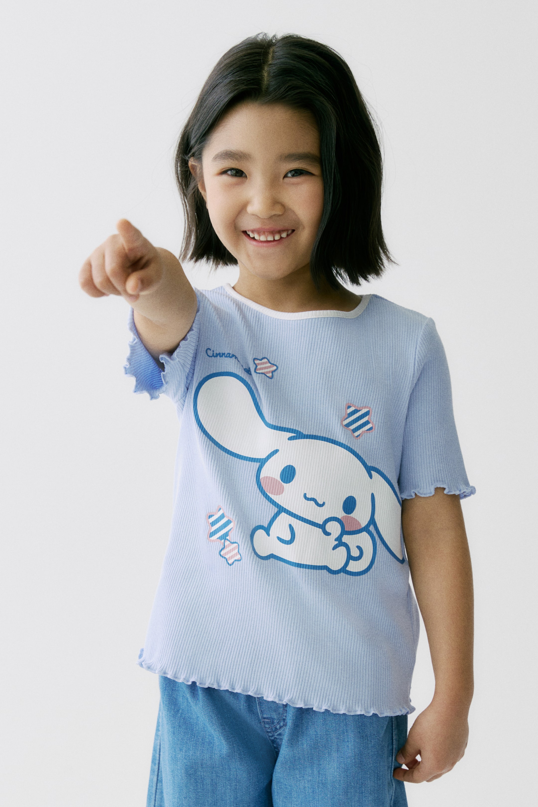 Voir une image plus grande: Un t-shirt côtelé bleu clair présente un grand imprimé du personnage Cinnamoroll en blanc et bleu sur le devant, accompagné du texte "Cinnamoroll" et de formes d'étoiles. Il est conçu avec des manches courtes à volants, un ourlet inférieur à volants et un col rond bordé de blanc, et est porté face avant sur un pantalon en denim bleu.