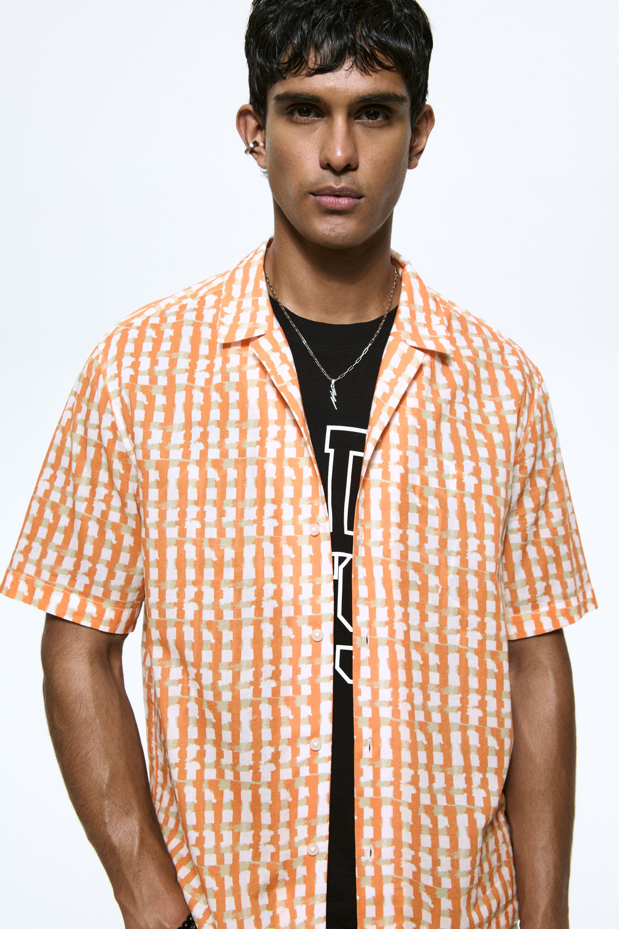 View larger image: An orange and off-white short-sleeve shirt with a brushed check pattern is worn open over a black T-shirt, facing forward. It features a camp collar, a button-down front with white buttons, and a single patch pocket on the left chest.