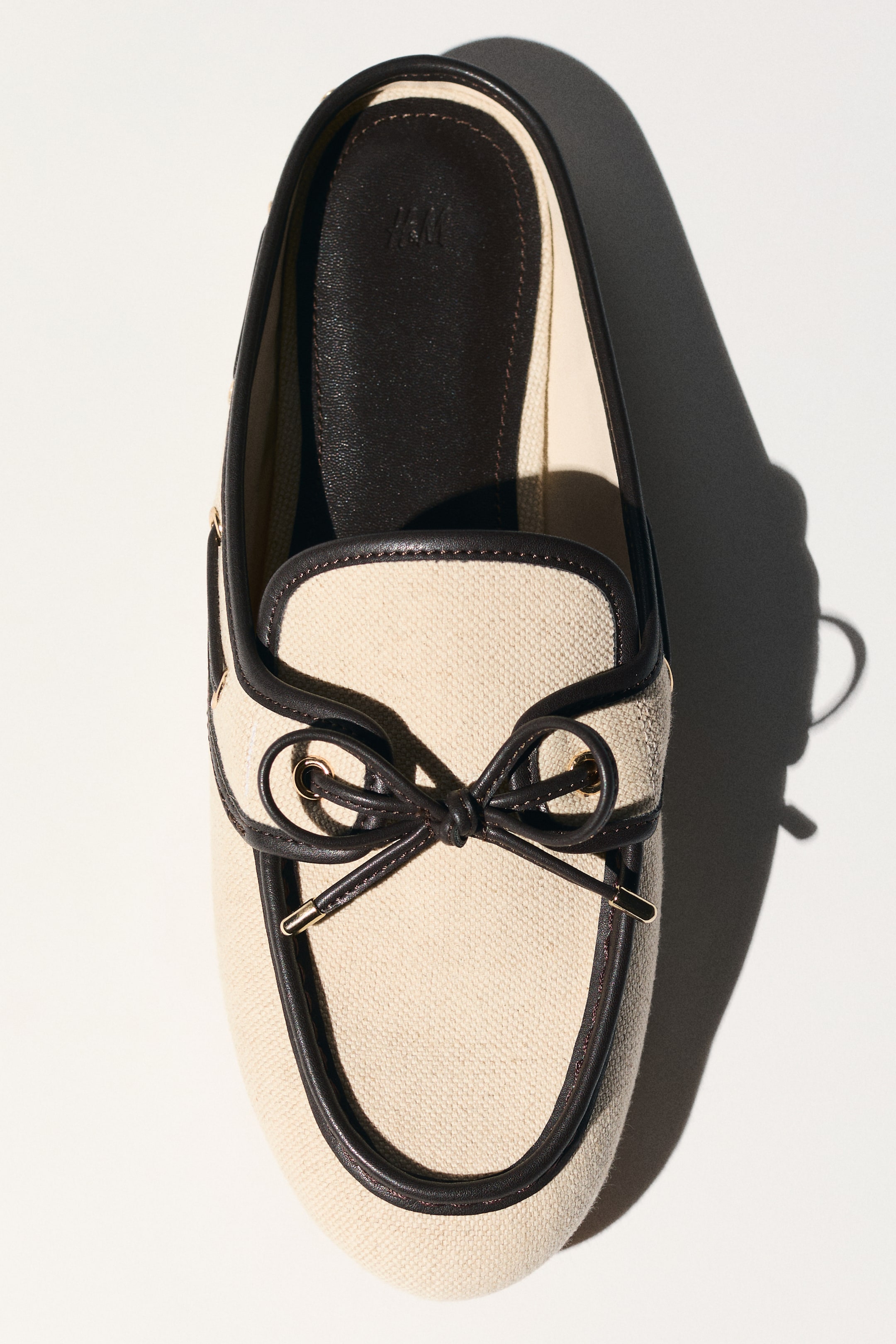 View larger image: A cream canvas mule faces forward, showcasing dark brown leather-like trim that outlines the shoe's opening, complemented by a decorative bow on the vamp. Gold-toned eyelets and aglets accentuate the bow, while a dark brown insole is visible within the backless design.