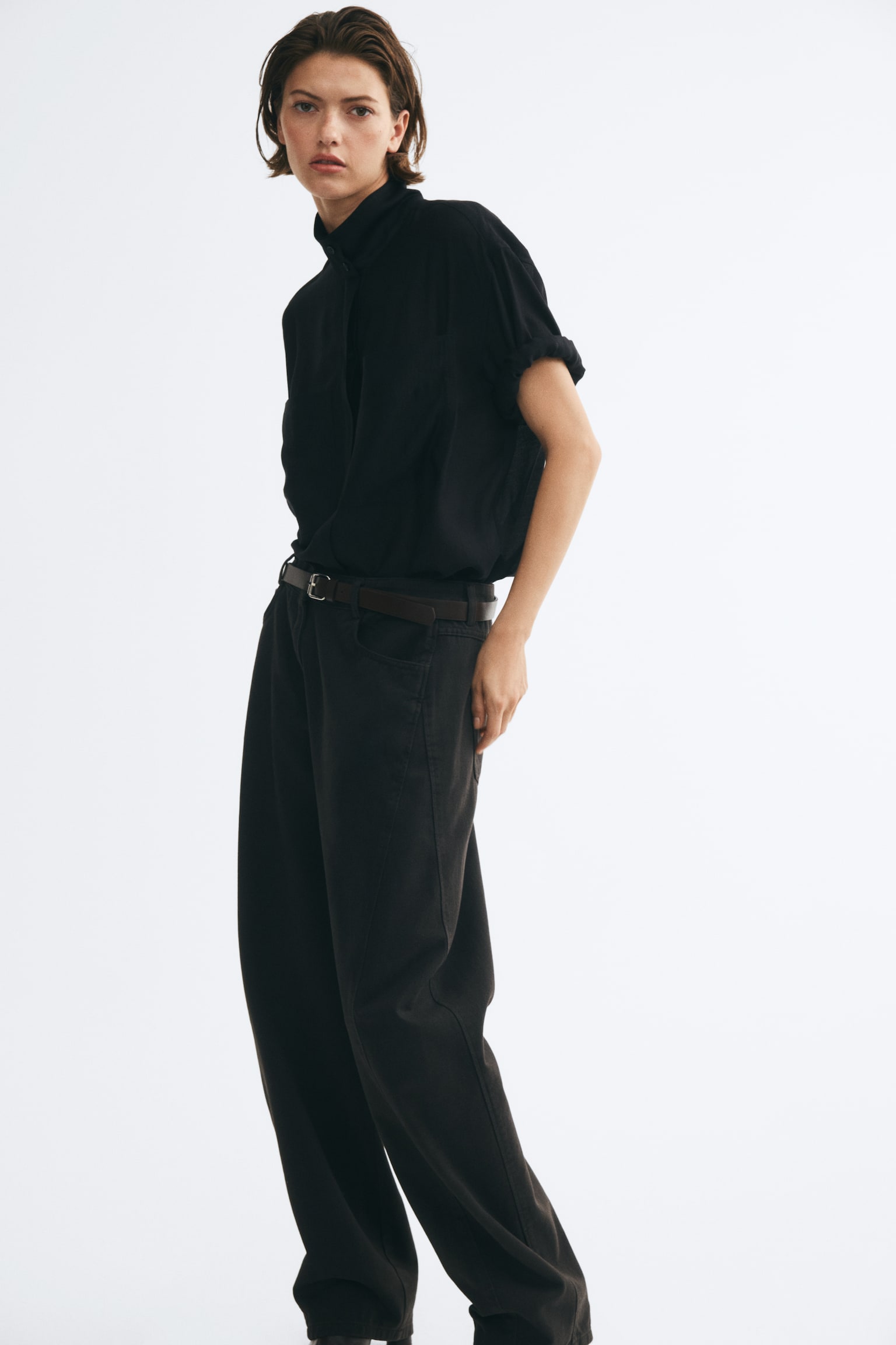 High-waisted, dark brown denim jeans are shown from the front, featuring a curved, voluminous leg silhouette that tapers towards the hem. The jeans are styled with a slim, matching brown belt with a silver buckle and feature prominent curved seams running down the sides. - 1