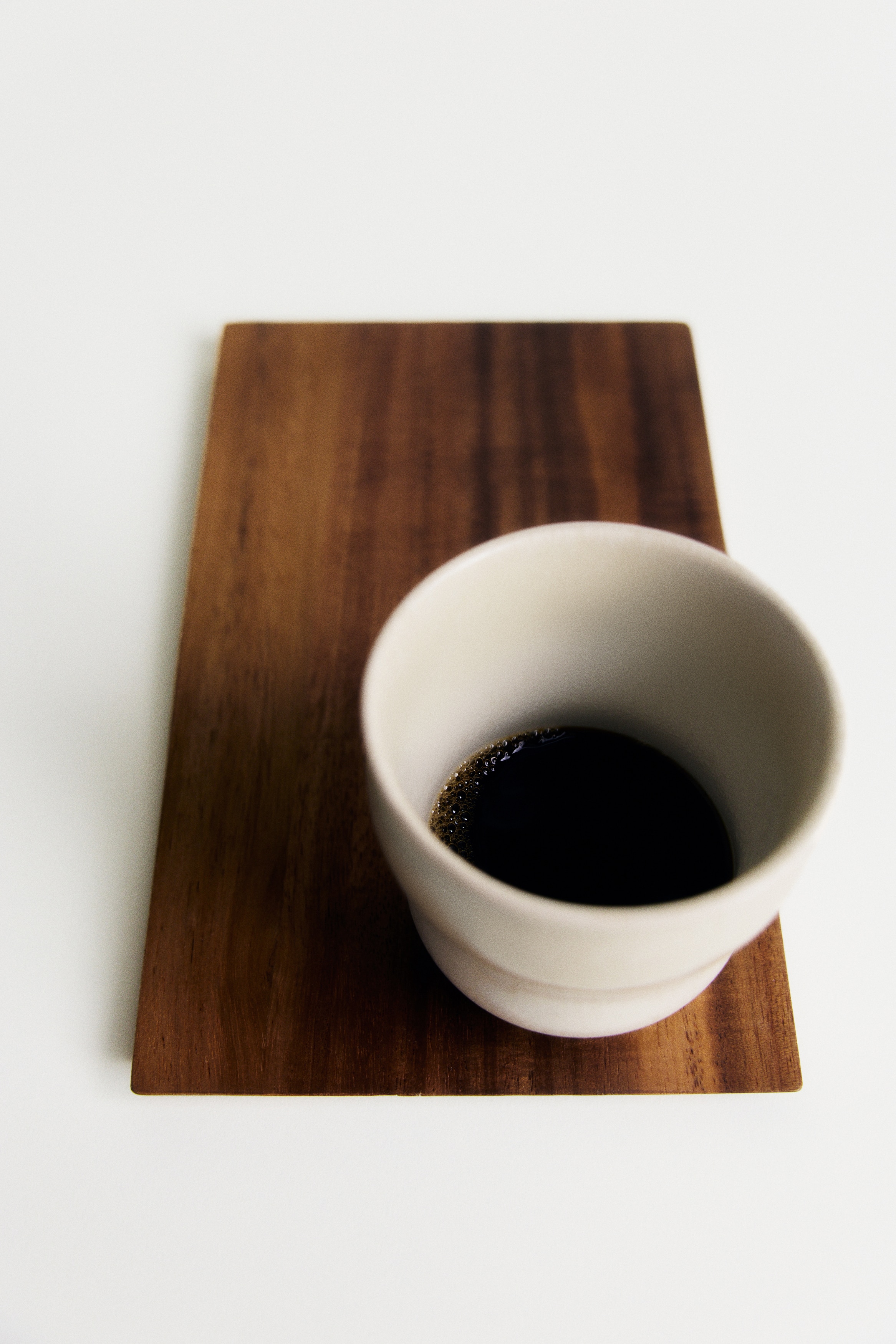 A gift set is featuring an off-white stoneware mug with a reactive glaze, displaying an irregular shape and subtle color variations, resting on a rectangular acacia wood tray. A light-colored gift box is standing behind the set, showing text on its front that reads 'FOR THE HOME BARISTA' and 'MUG & TRAY SET'.