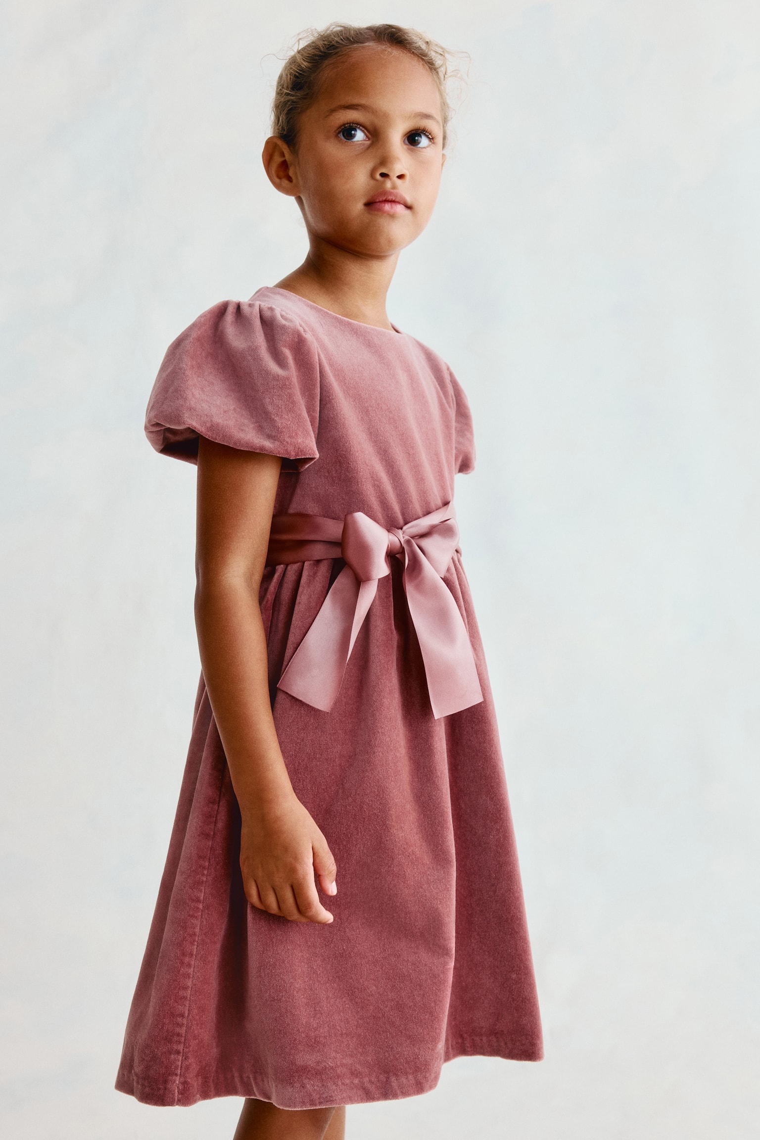 A dusty rose velvet dress is worn by a child facing three-quarters to the right, highlighting its round neckline and short puff sleeves. The dress features a matching satin sash tied in a bow at the waist and falls into a flared skirt. - 1
