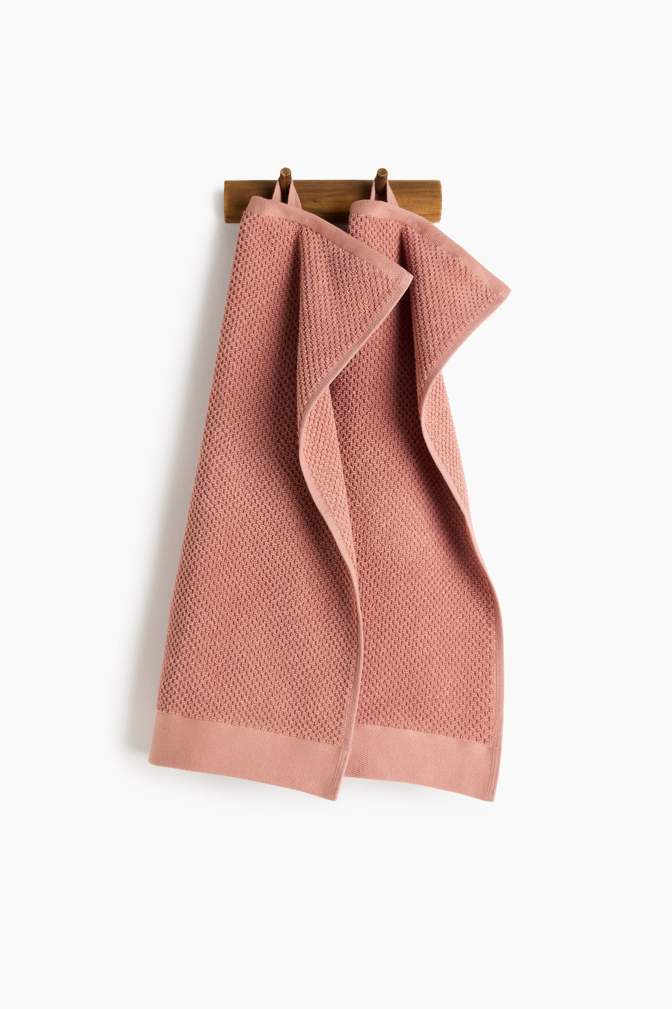 View larger image: Two pink guest towels are elegantly draped side-by-side on a wooden rack. Each towel features a textured pattern, a flat border, and a prominent loop at the top for easy hanging.