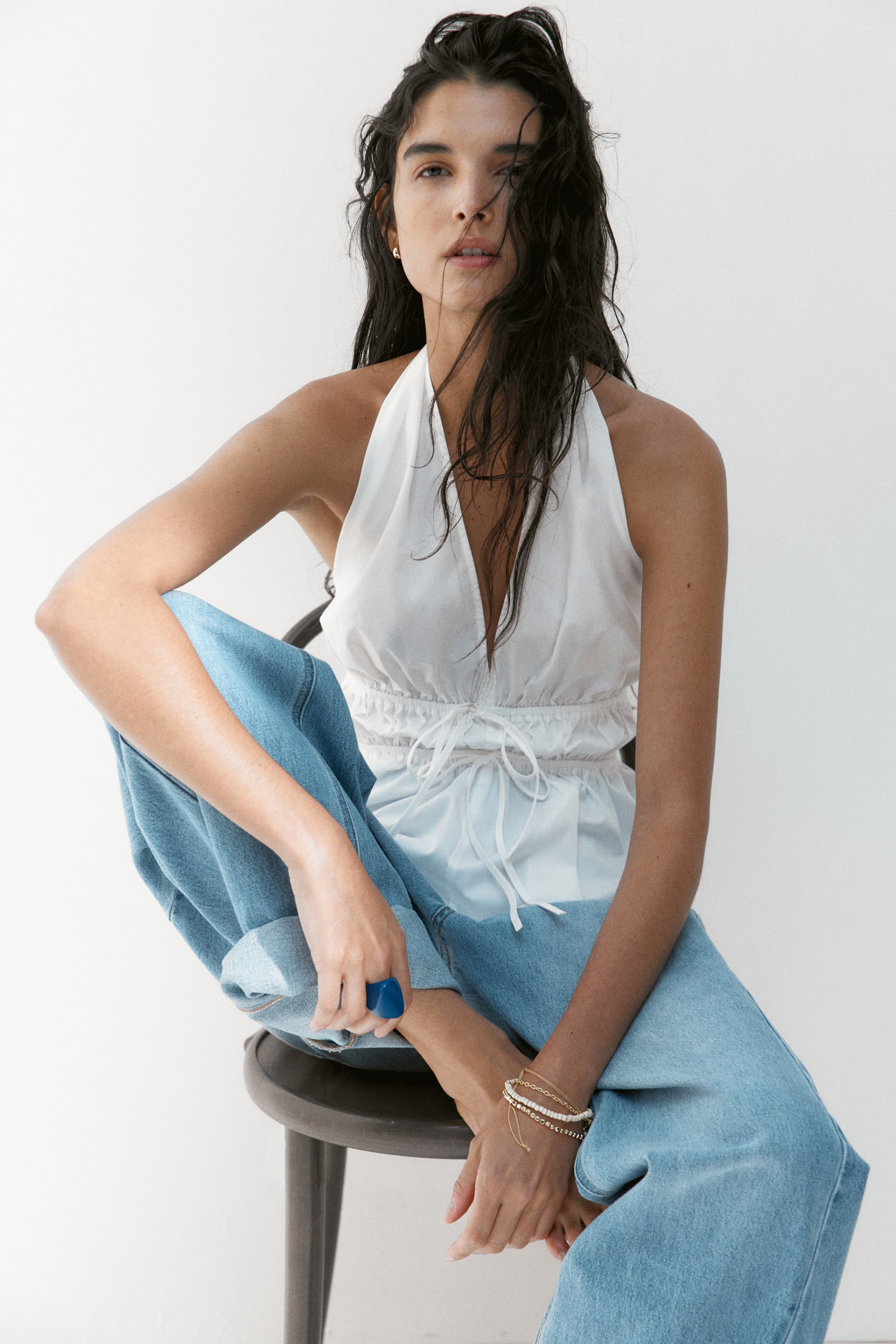 View larger image: A white halter neck top features a deep V-neckline and a cinched drawstring waist, with the strings tied at the front. It's paired with light blue, wide-leg jeans that are cuffed at the ankles.
