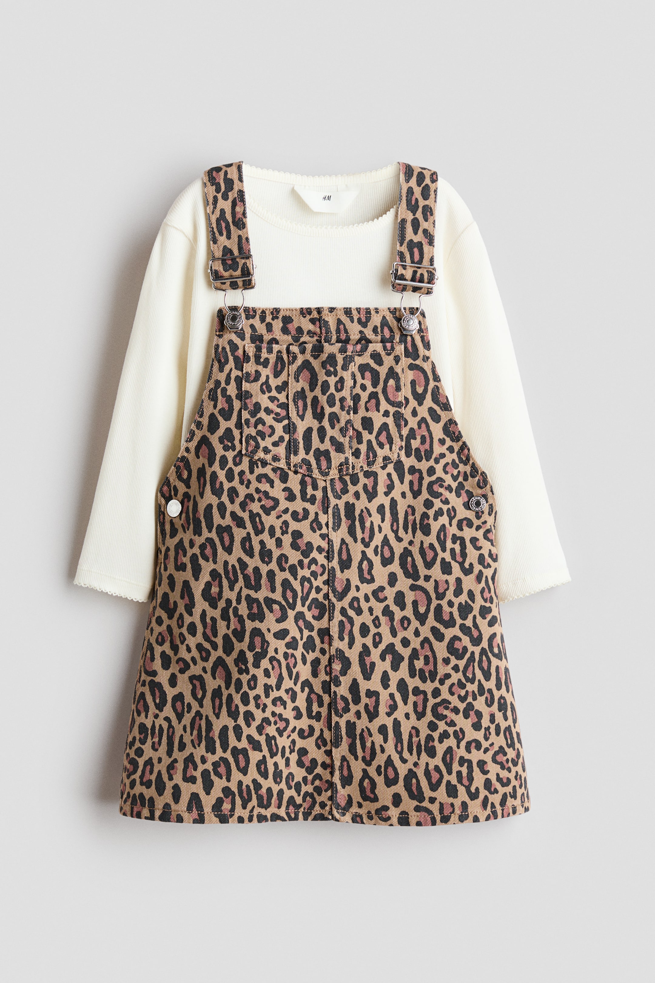 View larger image: A two-piece set showcasing an off-white, long-sleeved, ribbed jersey top with a picot-trimmed round neckline, layered under a leopard print denim dungaree dress. The dungaree dress features adjustable shoulder straps with metal fasteners, press-studs on the sides, and a chest pocket.