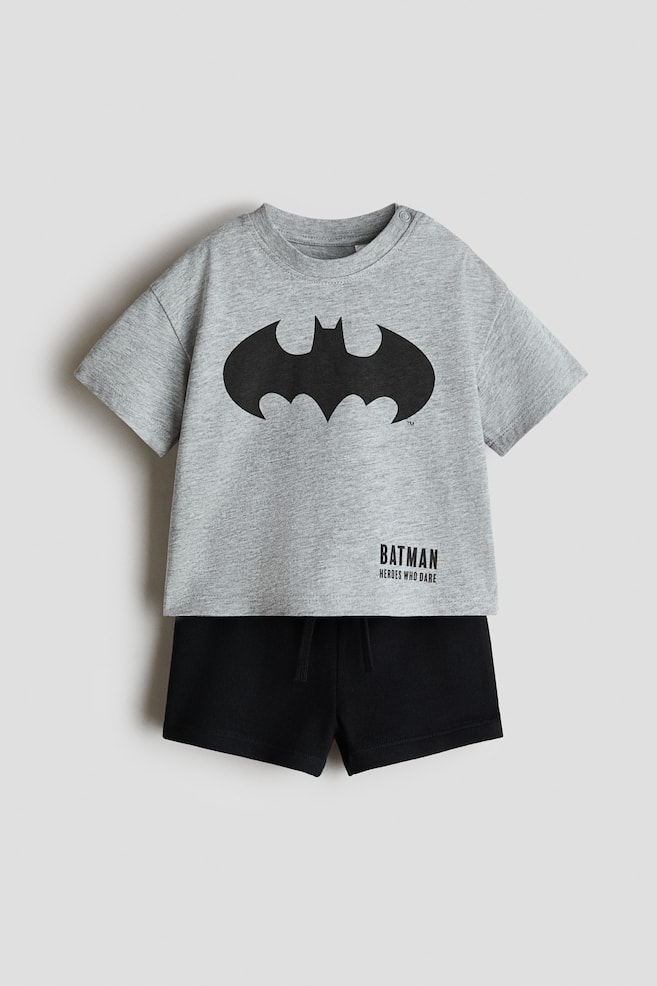 2-piece printed set, front-facing. A grey marl short-sleeved t-shirt is featuring a crew neck with snaps on one shoulder and a large black Batman logo on the chest, accompanied by "Batman Heroes Who Dare" text. Black shorts with an elasticated drawstring waistband are completing the set. - 1