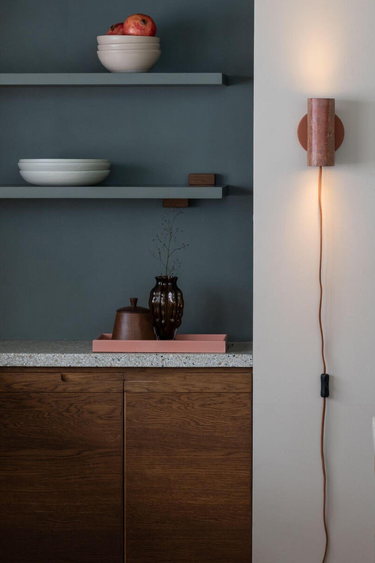 Deze wandlamp toont een verticale, cilindervormige kap van gestructureerde, roodbruine travertijnsteen met natuurlijke poriÃ«n. De stenen kap is bevestigd aan een matte, terracotta-kleurige, ronde wandbevestiging, met een bijpassend met stof omwikkeld snoer dat vanaf de onderzijde naar beneden loopt.