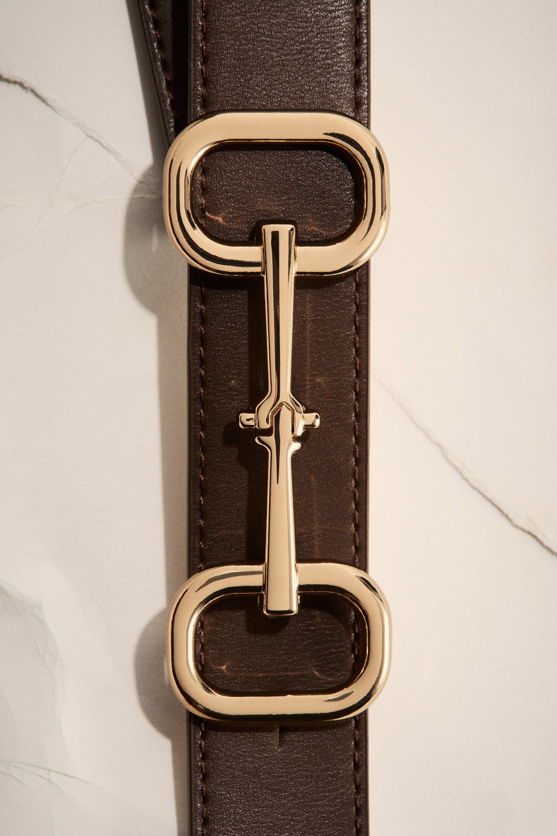 Ver imagem maior: Um cinto de couro castanho escuro apresenta uma peça de ferragem em estilo "horsebit" com acabamento dourado. Esta ferragem é composta por dois laços retangulares arredondados conectados por uma barra central com uma barra transversal decorativa menor, e a tira de couro exibe costuras visíveis ao longo das suas bordas.