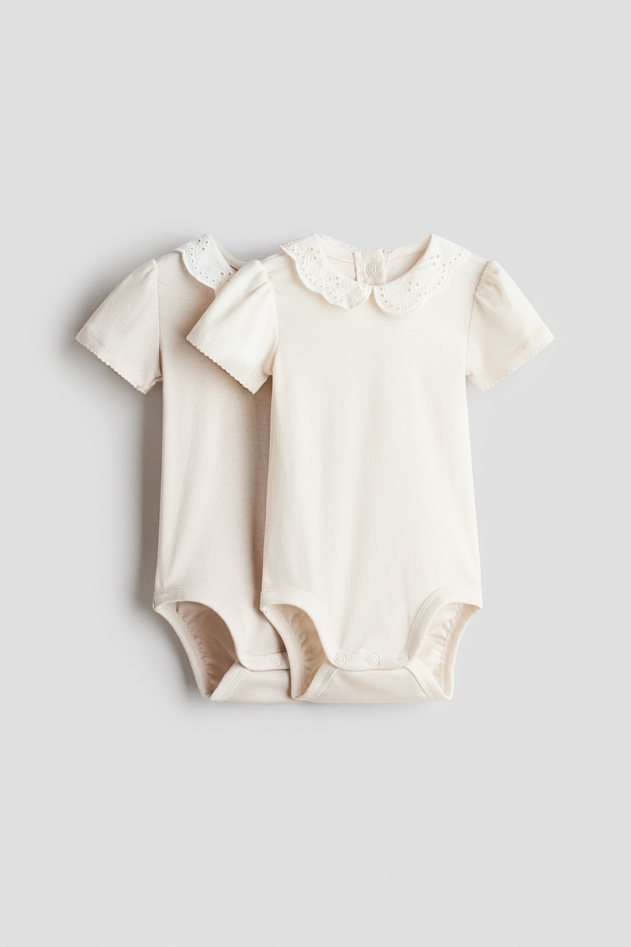 View larger image: Two cream-coloured baby bodysuits feature short puffed sleeves, an embroidered scalloped Peter Pan collar, snap fasteners at the back of the neck, and snap fasteners at the crotch.
