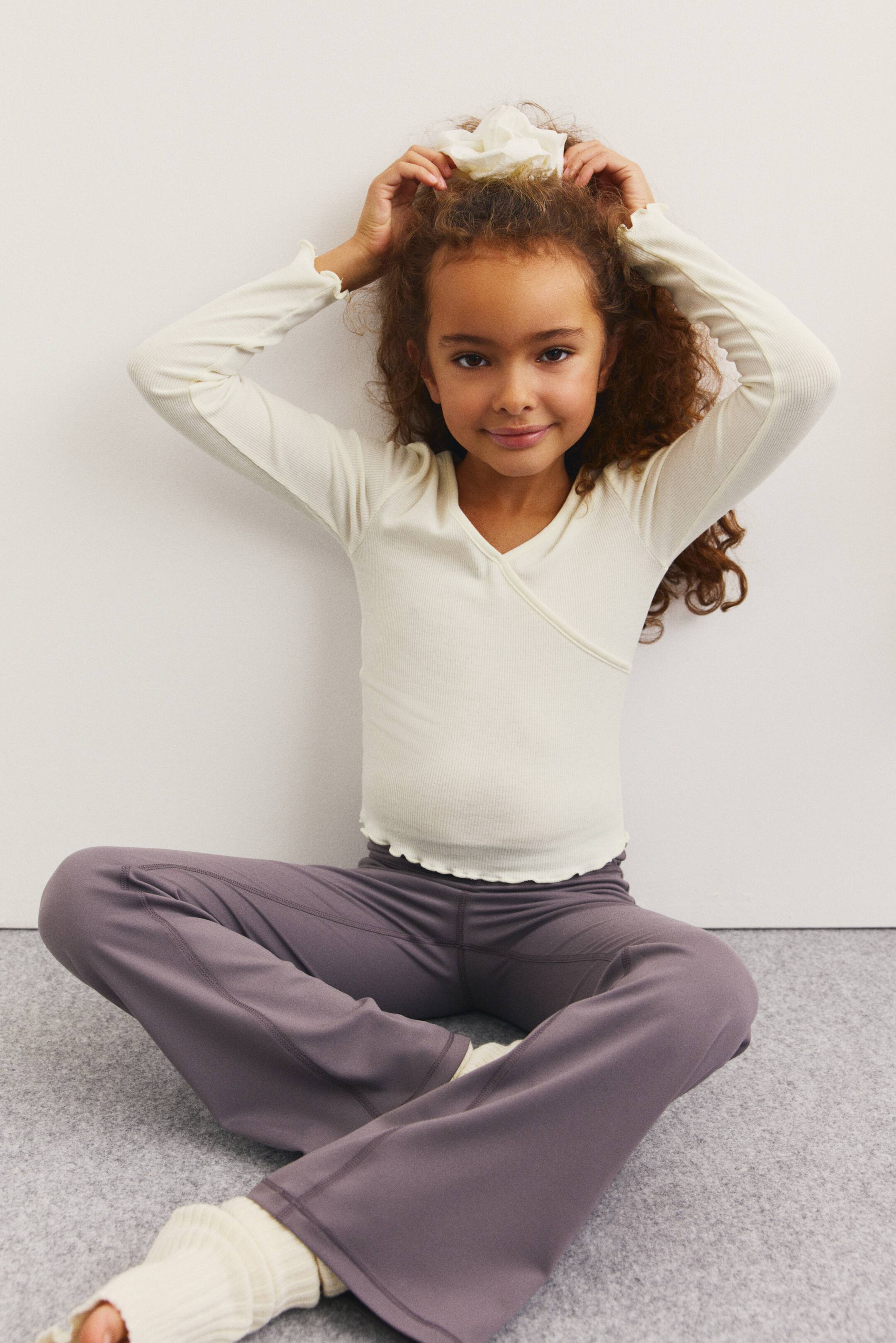 Ampliar la imagen: Se presenta un top color crema acanalado, envolvente, de manga larga, con escote en V y puños y dobladillo con volantes, visto desde un ángulo frontal que muestra su ajuste. Se usa con pantalones deportivos color morado oscuro con costuras visibles, y sobre los pantalones, a la altura de los tobillos, se llevan calentadores acanalados color crema.