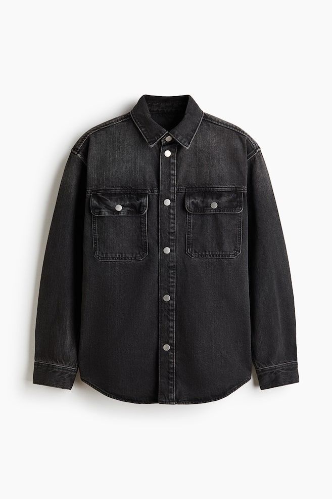 Ein gewaschenes schwarzes Denim-Hemd präsentiert seine Vorderansicht und zeigt einen klassischen spitzen Kragen sowie eine vollständige Druckknopfleiste mit silberfarbenen Knöpfen. Zwei Brusttaschen mit Patten und Druckknopfverschlüssen sind sichtbar, zusammen mit langen Ärmeln, die in Manschetten mit einem einzelnen Druckknopf enden, und einem abgerundeten Saum. - 1
