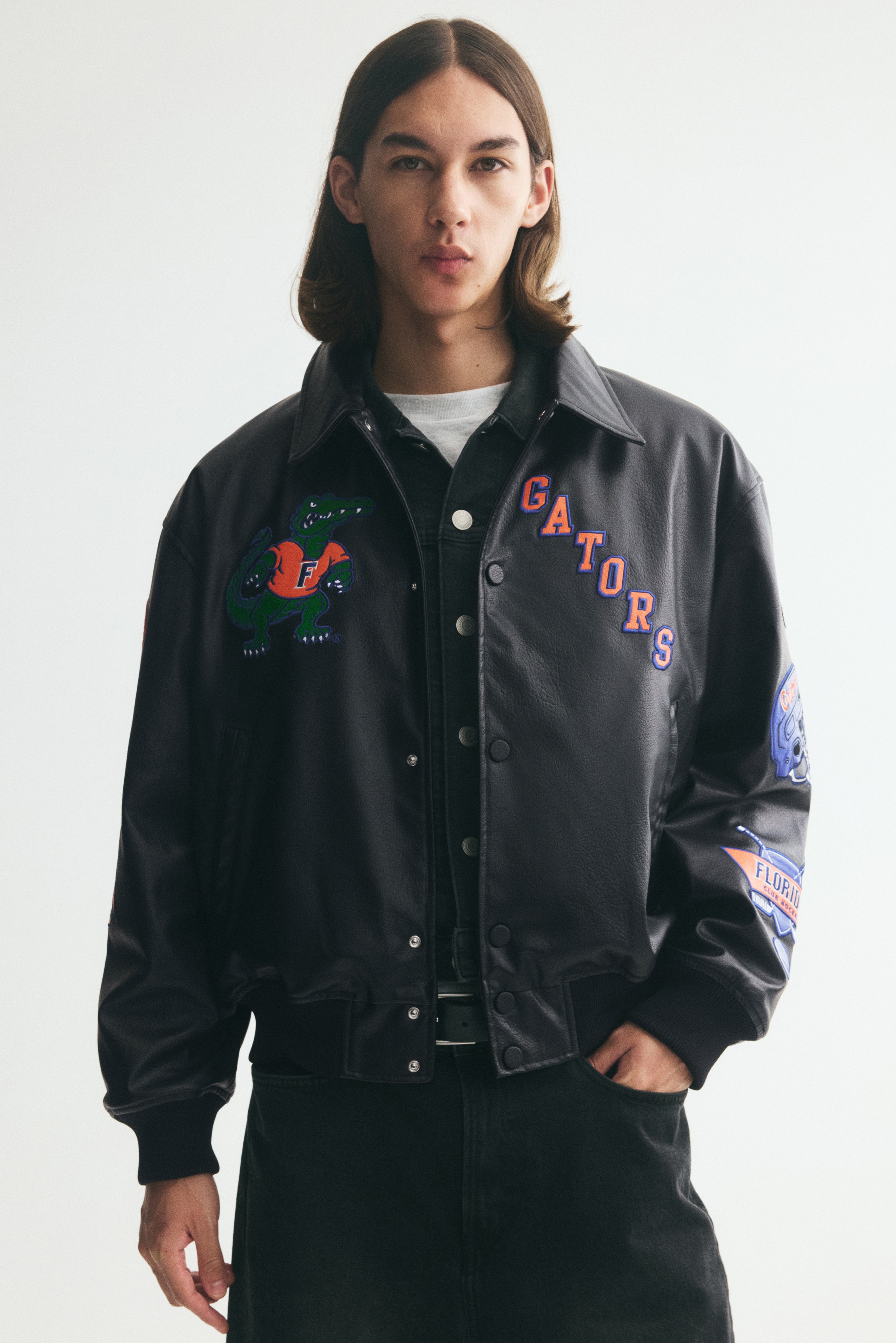 A black faux-leather jacket is shown from the front, featuring a pointed collar, snap-button front closure, slanted welt pockets, and ribbed cuffs and hem. The jacket is decorated with colourful sports-themed patches, including a green alligator mascot on the right chest, the word "GATORS" on the left chest, and various graphic emblems along both sleeves.