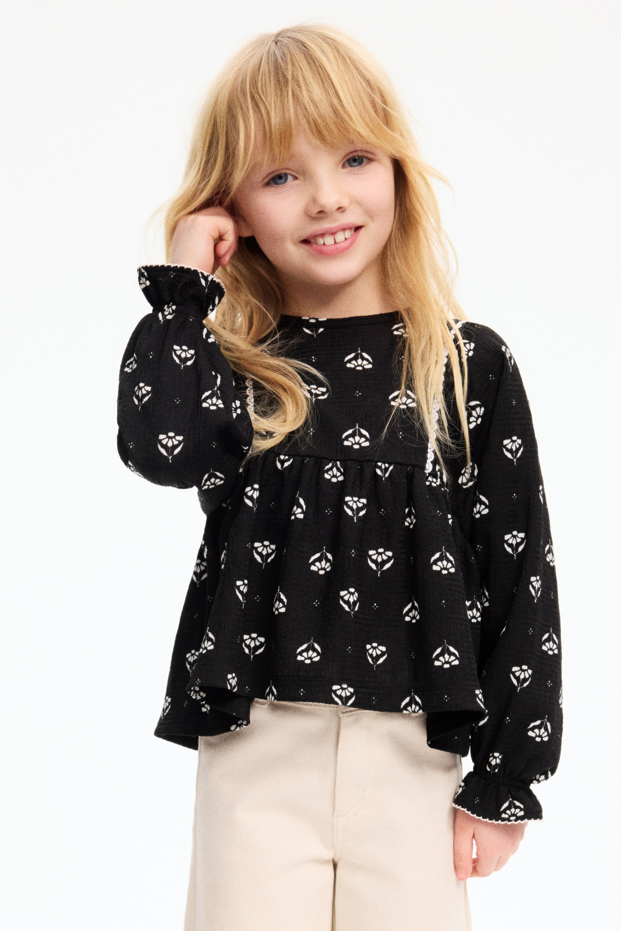 이미지 확대 보기: 블랙 컬러의 긴소매 탑으로, 텍스처가 느껴지는 원단에 화이트로 스타일화된 플로럴 및 도트 패턴이 들어가 있습니다. 라운드 네크라인, 셔링이 잡힌 소매, 화이트 트림의 러플 커프스로 디자인되어 여유롭고 흐르는 듯한 핏을 연출합니다.