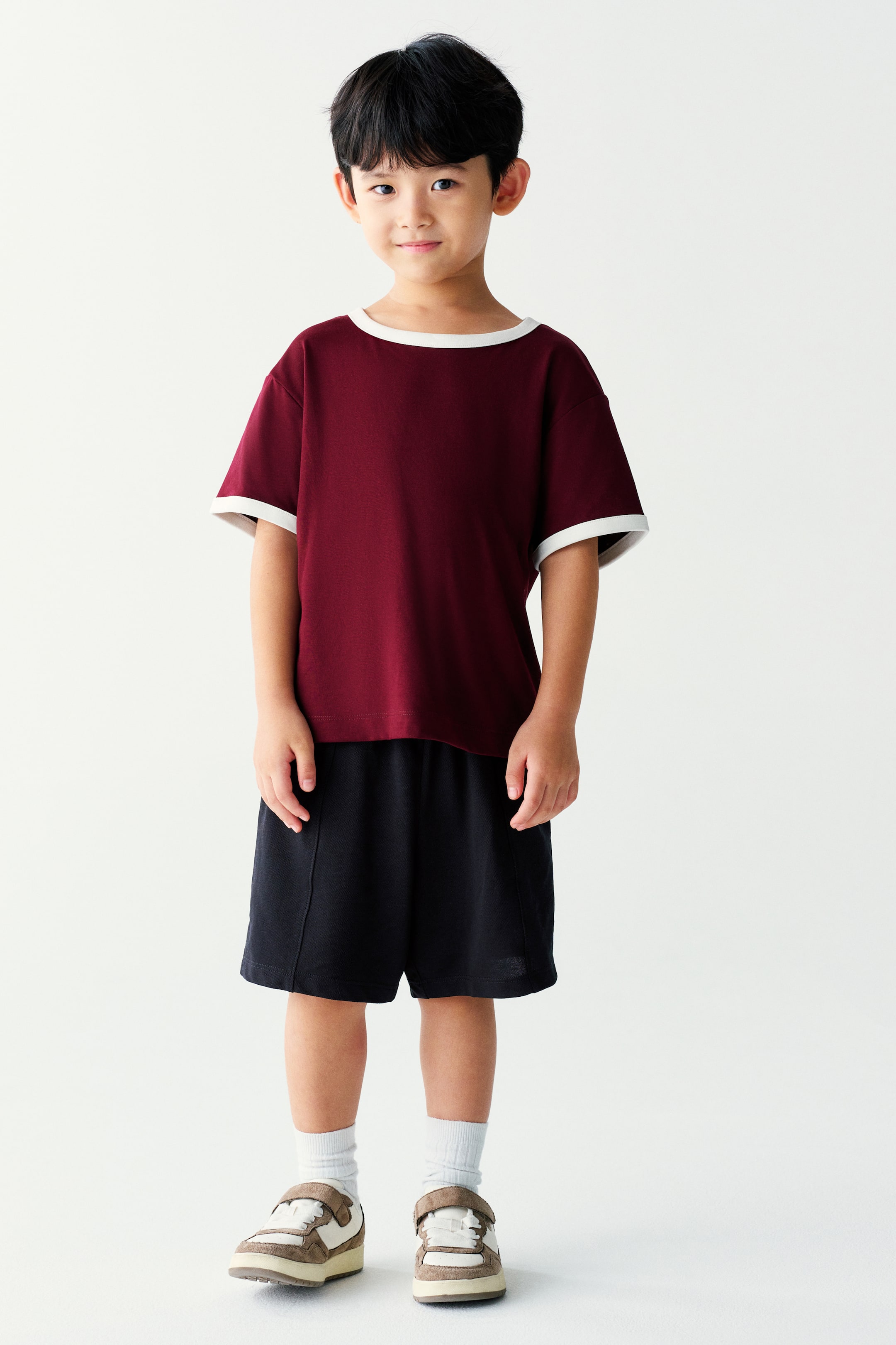 Xem ảnh lớn hơn: Một chiếc áo thun tay ngắn màu đỏ burgundy với cổ tròn tương phản màu trắng và bo tay áo đồng màu đang được người mẫu mặc cùng quần short đen dài đến giữa đùi. Bộ trang phục casual được phối với tất trắng và giày sneaker cổ thấp.