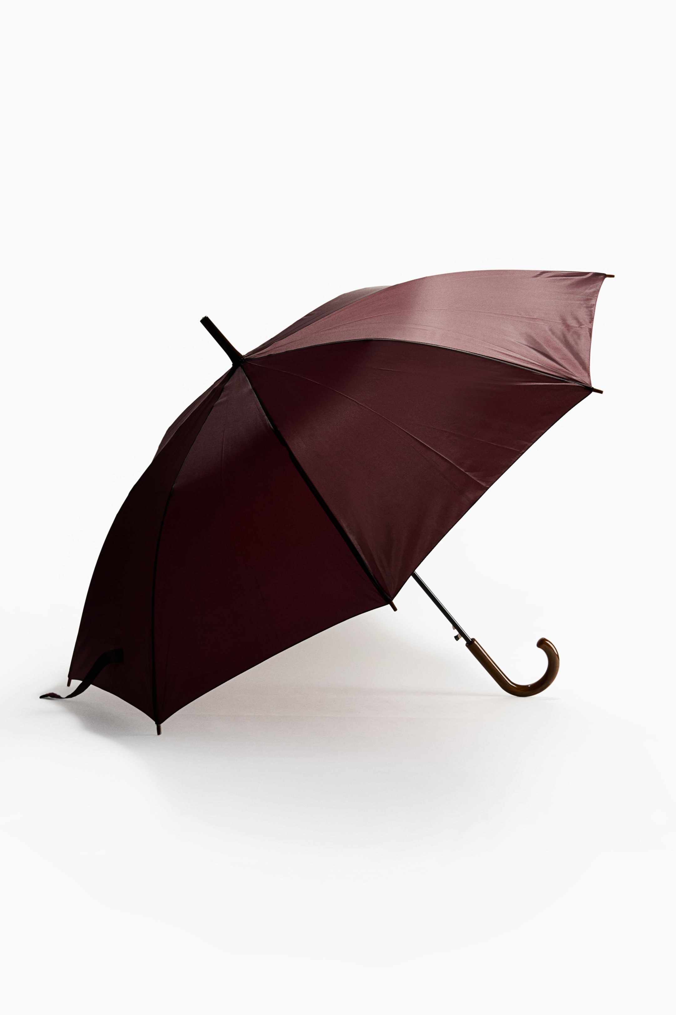 Vezi imaginea mai mare: O umbrelă burgundy deschisă este sprijinită pe o parte, afișând copertina sa maro-închis și un mâner curbat din lemn, tip baston. Tija și vârful metalice negre sunt vizibile în timp ce umbrela este înclinată.