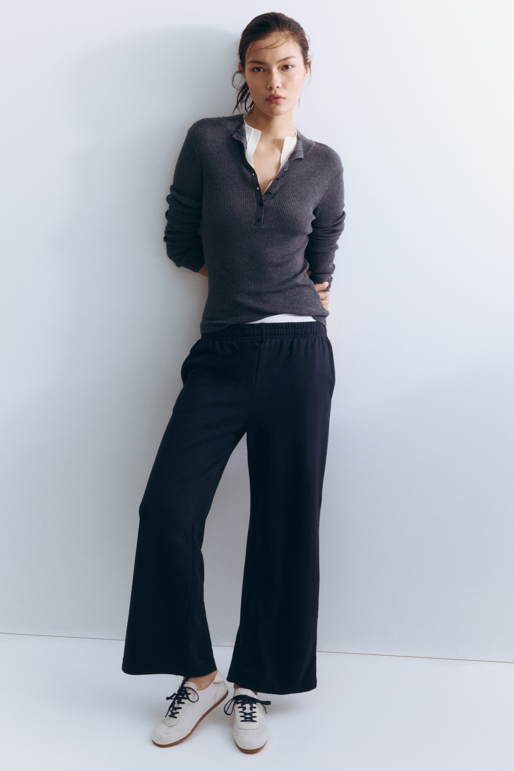 View larger image: From a front-facing perspective, a dark gray ribbed long-sleeved polo-style top is fitting snugly, featuring a collared neckline, a three-button placket, and a white undershirt visible at the collar and hem. Black wide-leg pants are displaying an elastic waistband and a relaxed fit, while cream-colored sneakers are also visible, featuring dark laces and light brown soles.