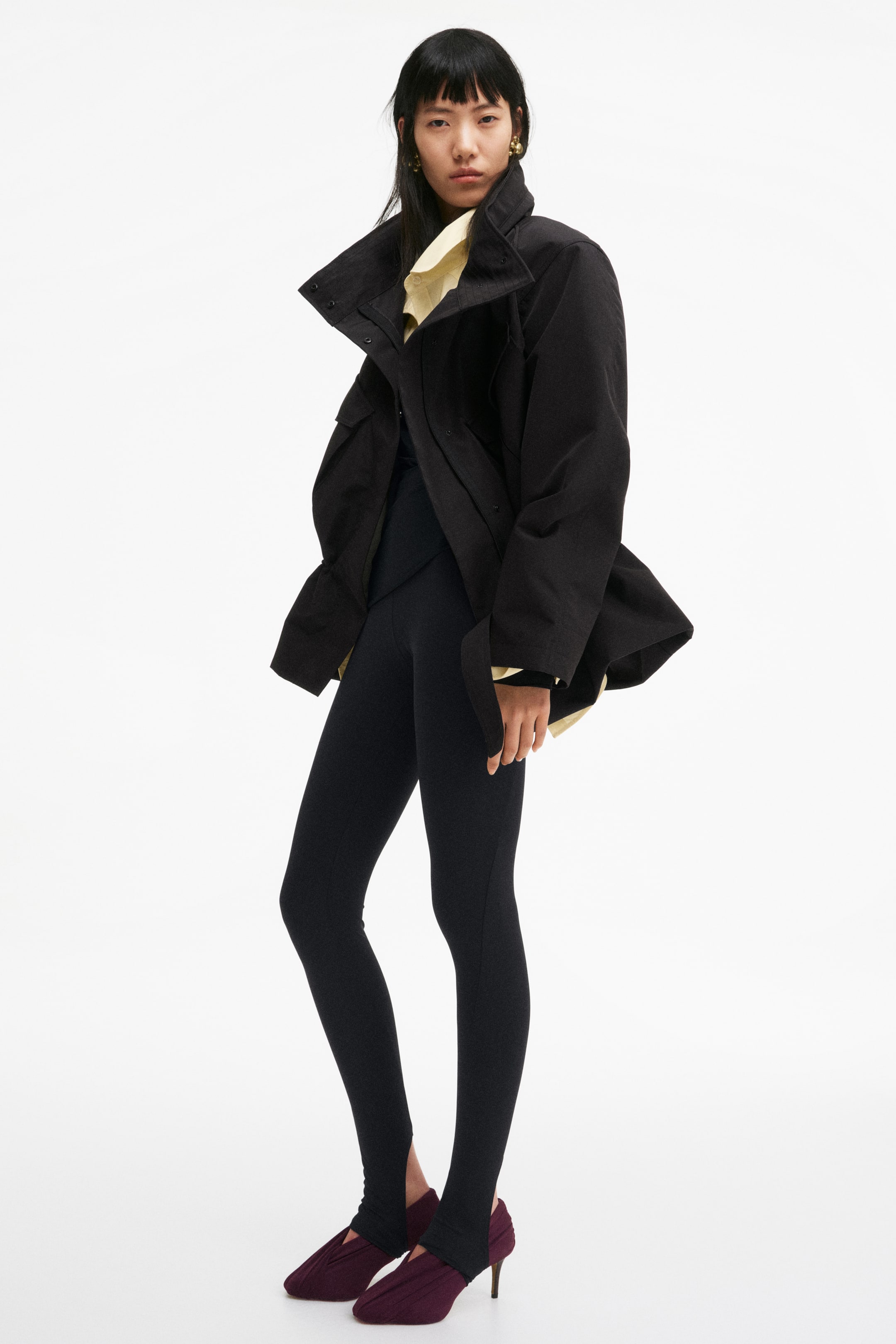 View larger image: An oversized black jacket features a high, structured collar and wide sleeves, worn open over a light yellow top. Black form-fitting stirrup leggings extend over burgundy pleated, heeled shoes.