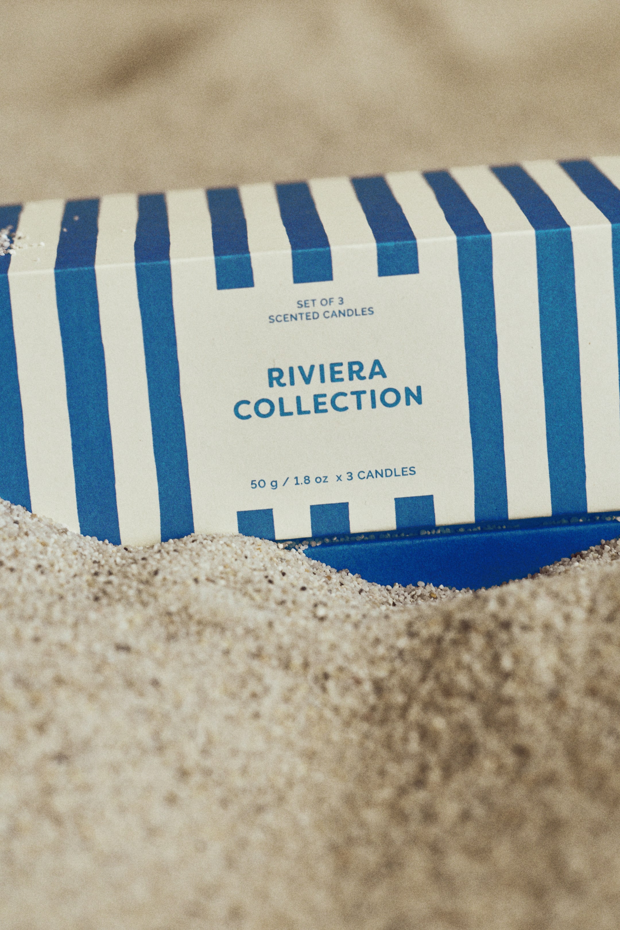 Se større billede: Et gavesæt præsenteres i en frontvendt, blå- og hvidstribet papæske. Æsken viser teksten "SÆT MED 3 DUFTLYS," "RIVIERA COLLECTION," og "50 g / 1,8 oz x 3 LYS."