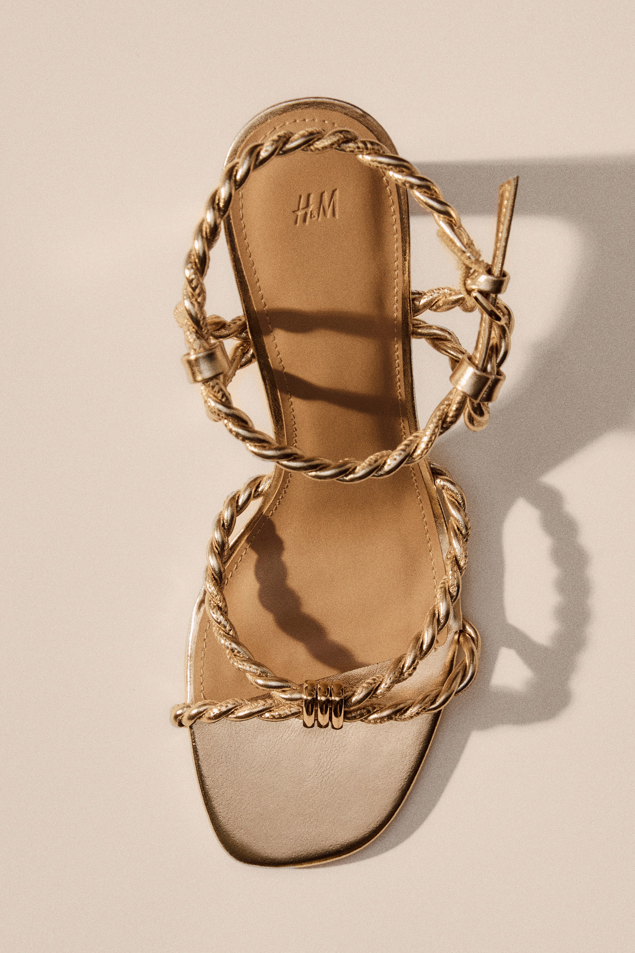Größeres Bild anzeigen: Goldfarbene Sandalen mit beschichtetem Stoff, verdrehten Fußriemen und einem Knöchelriemen mit Schnalle auf einer Seite, präsentieren sich mit quadratischen Zehen und einem hellbraunen Fußbett mit H&M-Logo.