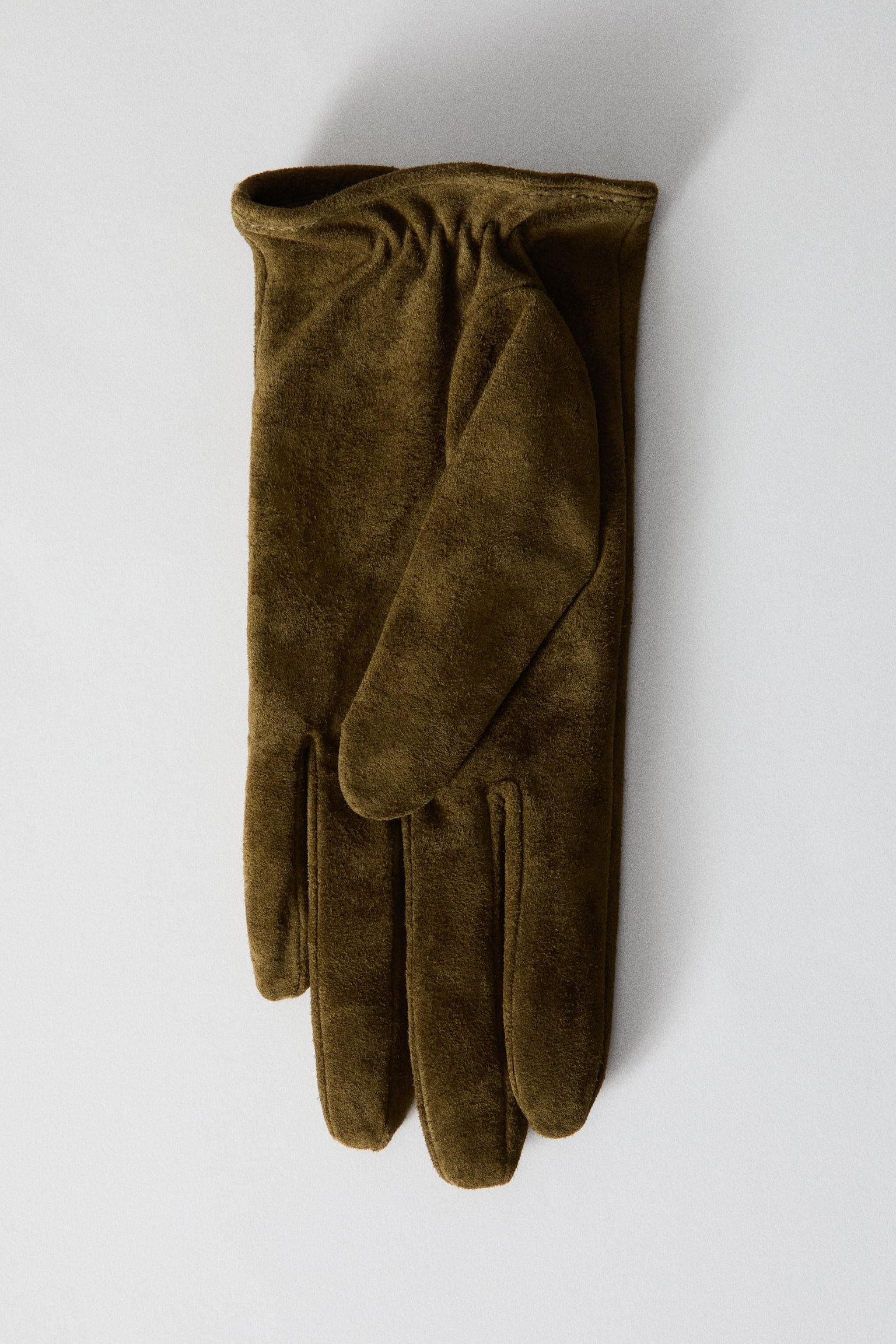 A pair of olive-green suede gloves is lying flat, showing their soft, matte texture. The glove at the front shows a gathered, elasticated wrist detail and a folded thumb. - 1